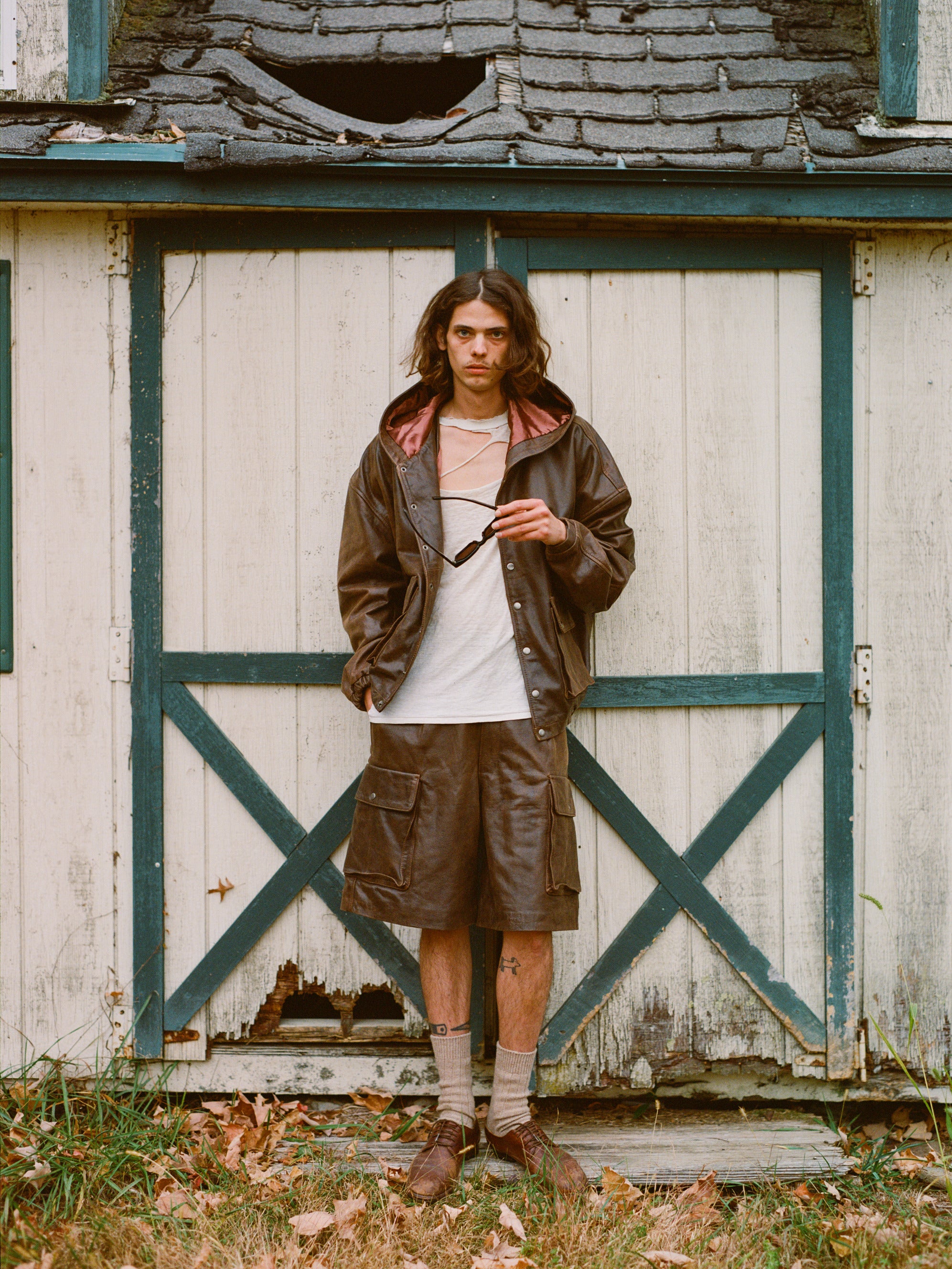 A person in casual, layered clothing, featuring Found's Dusk Distressed Genuine Leather Cargo Shorts, stands by a weathered wooden structure with green and white doors. With long hair and sunglasses in hand, they wear socks and shoes, embracing a utilitarian style that fuses fashion with function.