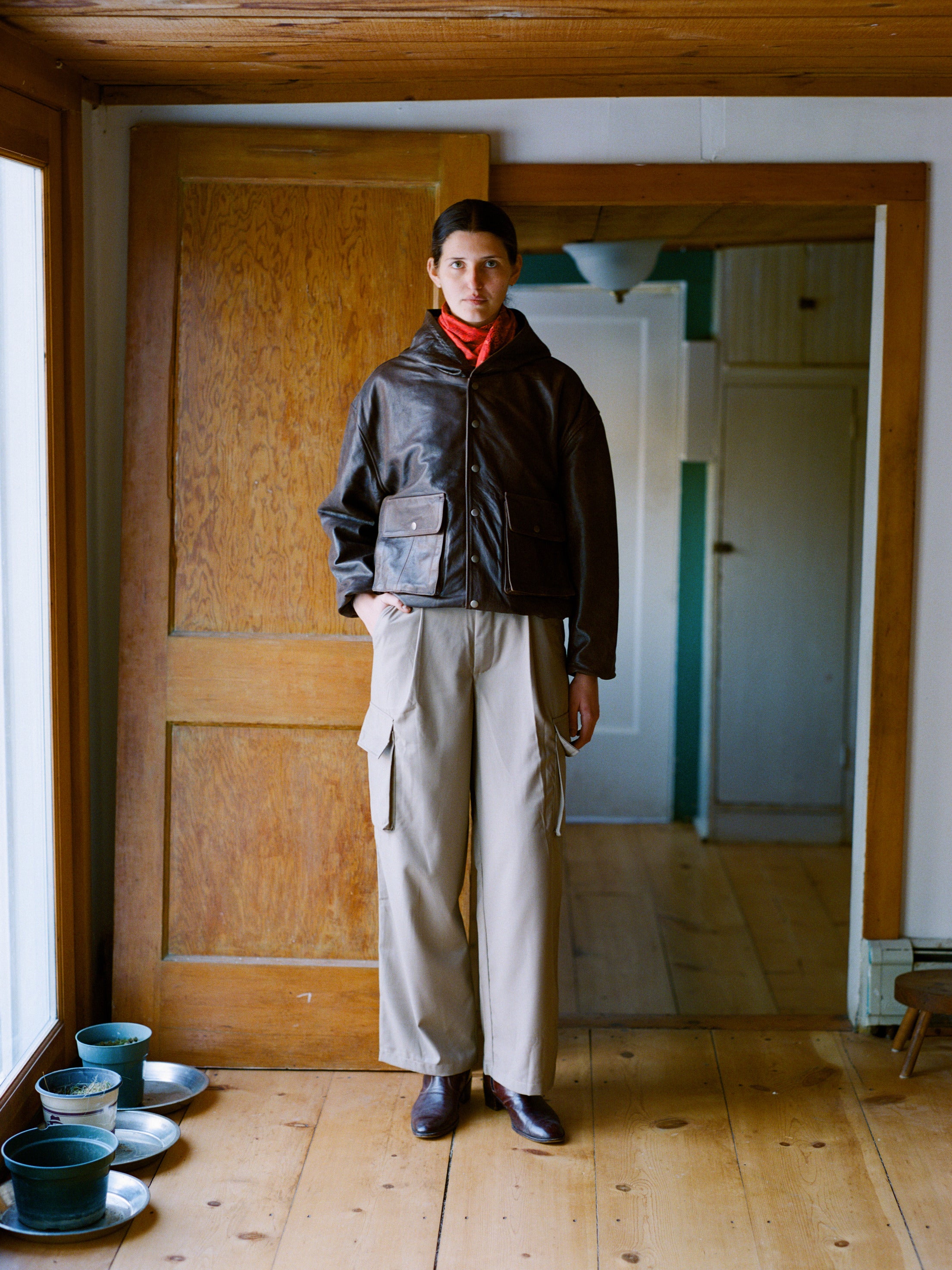 An individual dons a dark leather jacket, red scarf, and FOUND's Hava Pleated Cargo Trousers indoors, effortlessly embodying a utilitarian style against the backdrop of wooden floors and a door.