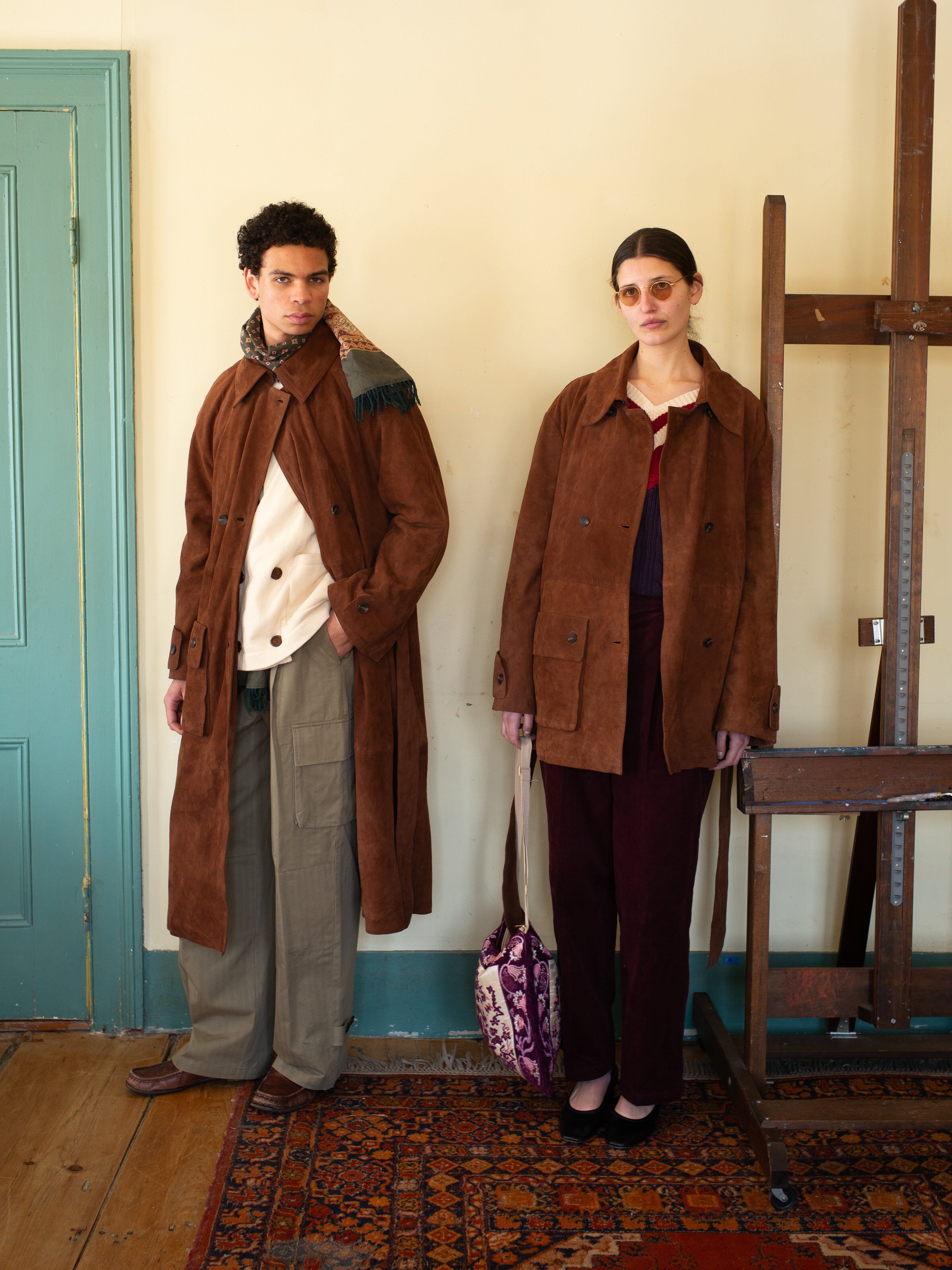 Two people in brown jackets stand indoors, showcasing utility wear. One carries a purple and white bag, posing by an easel on a patterned rug near a light green door. They're wearing FOUND's Hava Herringbone Pleated Cargo Trousers, blending wool-blend fabric style with function.