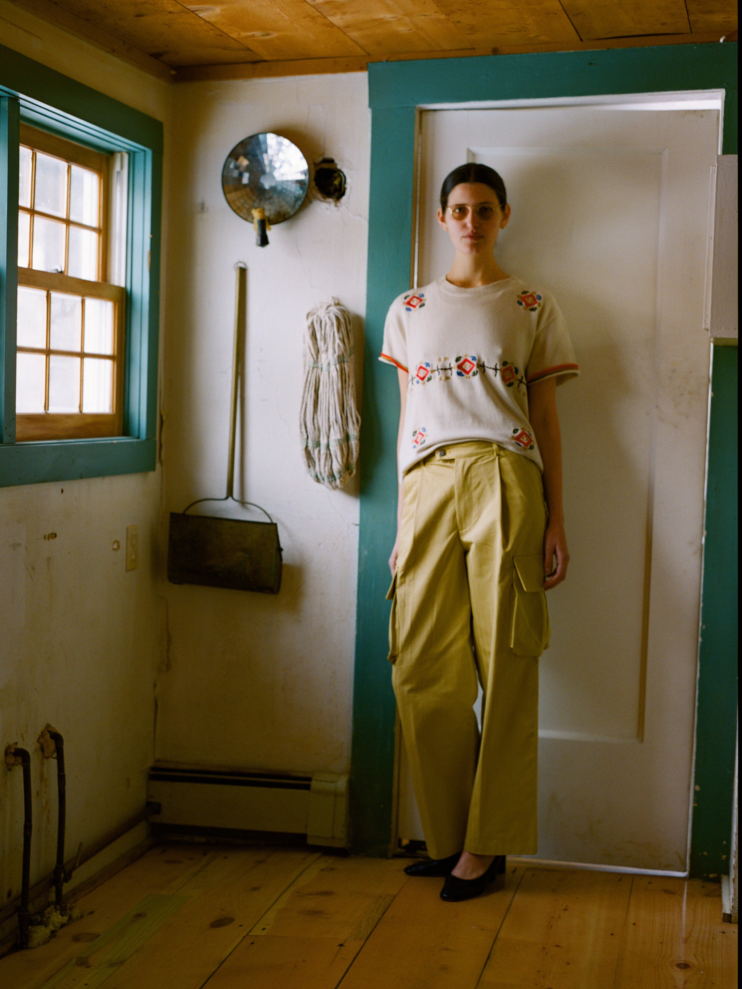 In a rustic room with a wooden floor, teal-trimmed window and door, and hanging tools on the wall, a person wears FOUND's Knit Flower Petal Tee paired with beige cargo pants, exuding vintage-inspired charm.