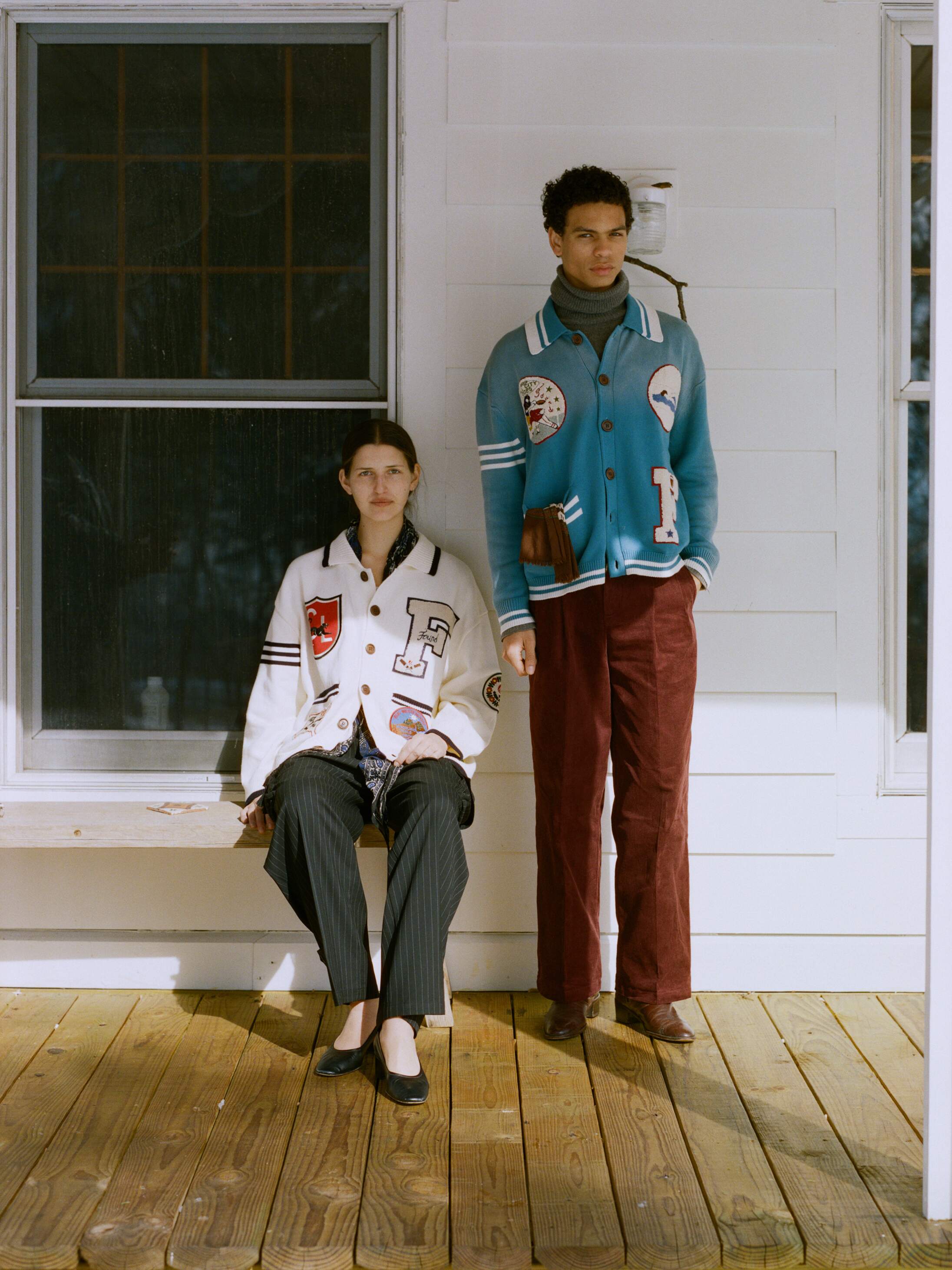 Two people are on a wooden porch. One sits in a letterman-style jacket and striped pants, while the other stands, showcasing collegiate style with brown pants and the FOUND Fin Varsity Patch Collared Cardigan.