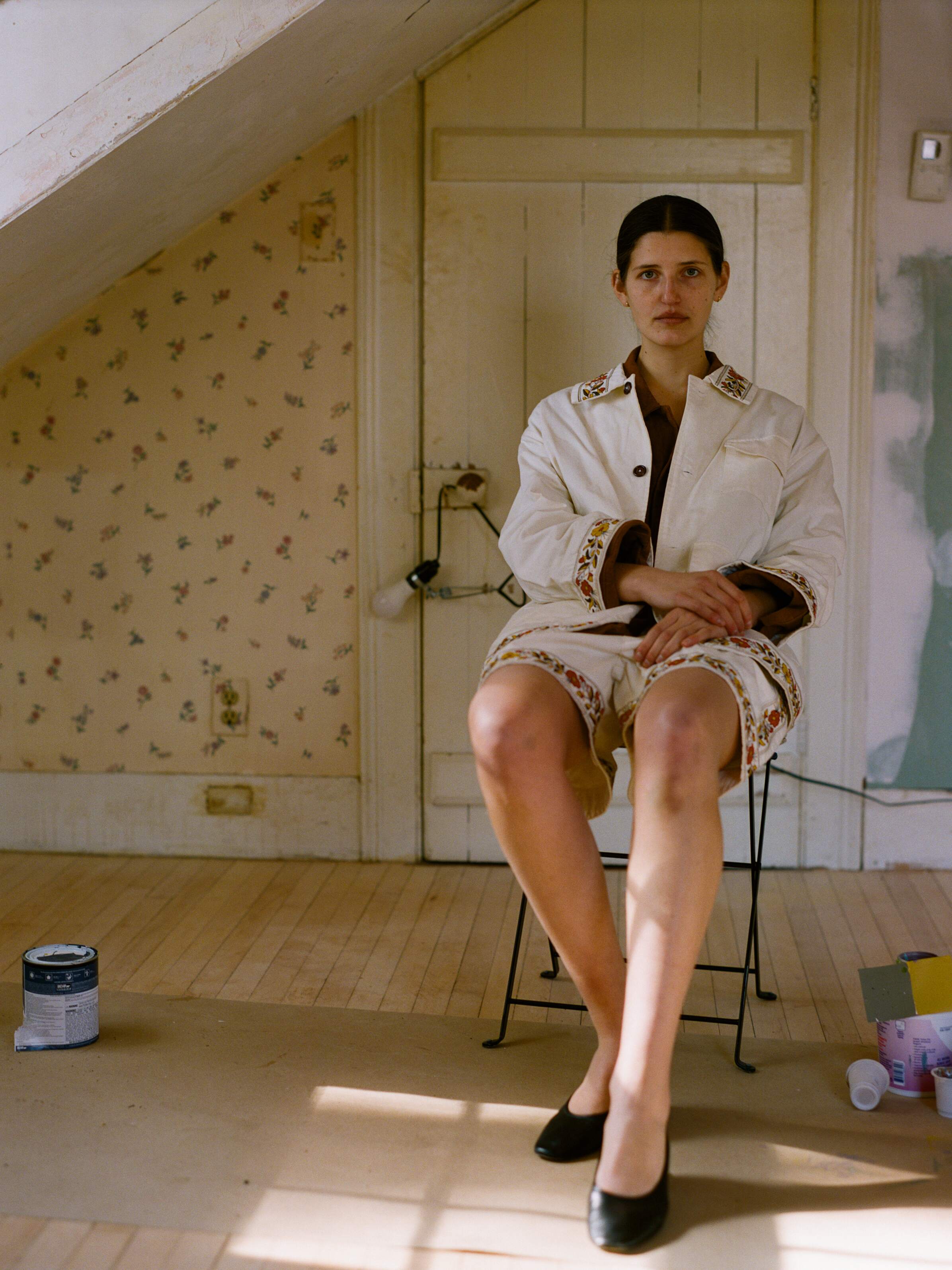 In a room with floral wallpaper and a slanted ceiling, someone in a white outfit with patterned trim and black shoes sits nearby. Paint cans lie on FOUND's 100% cotton Kantha Floral Work Shorts spread across the floor.