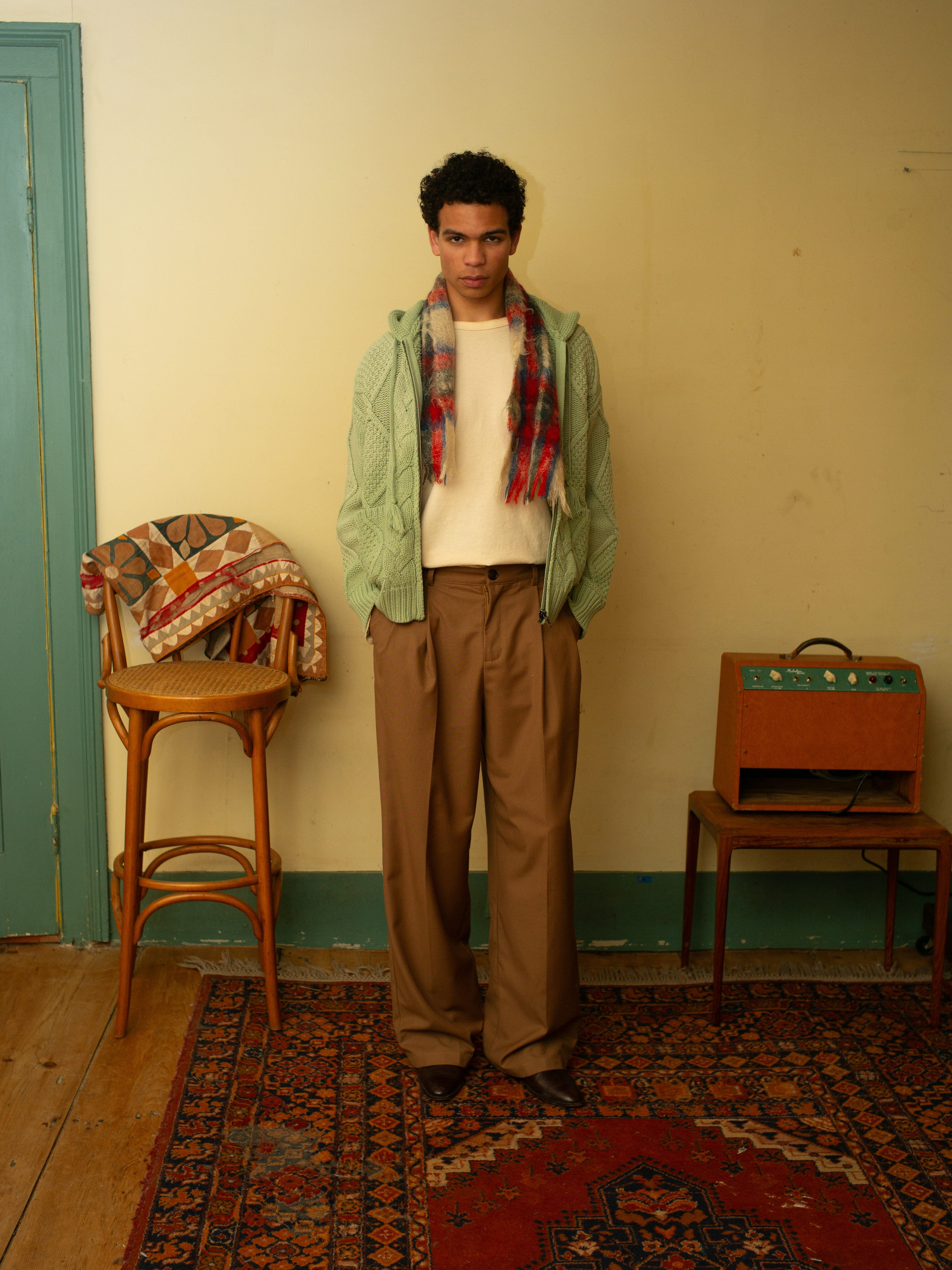 Person indoors in a relaxed-fit Zip Up Cable Knit Hoodie by Found, light green, layered over a white shirt and plaid scarf with brown pants. A patterned rug, fabric-covered chair, and wooden cabinet enhance the setting.