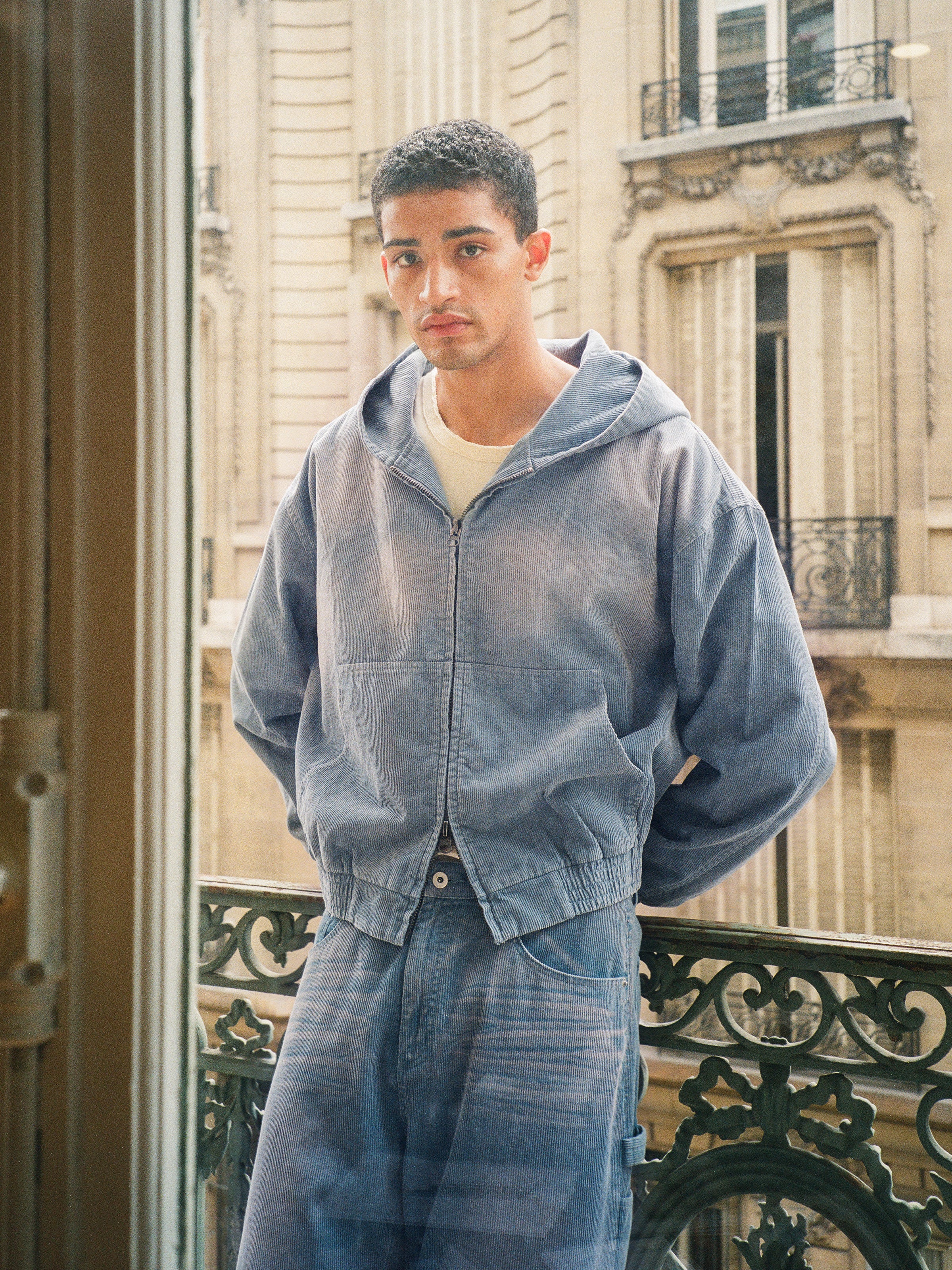 A person stands on a balcony wearing the FOUND Corduroy Zip Up Hoodie and jeans, with classic stone buildings and window railings in the background.