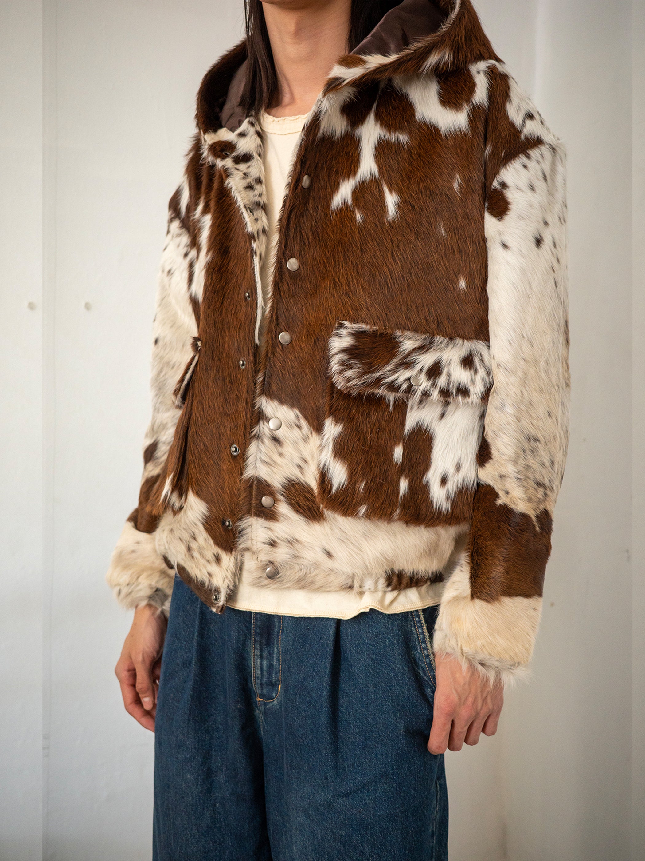 A person wears the FOUND Cowhide Drover Hoodie, featuring a brown and white cowhide pattern with snap buttons, paired with blue jeans and a cream shirt. The cropped frame focuses on this standout unisex outerwear piece.