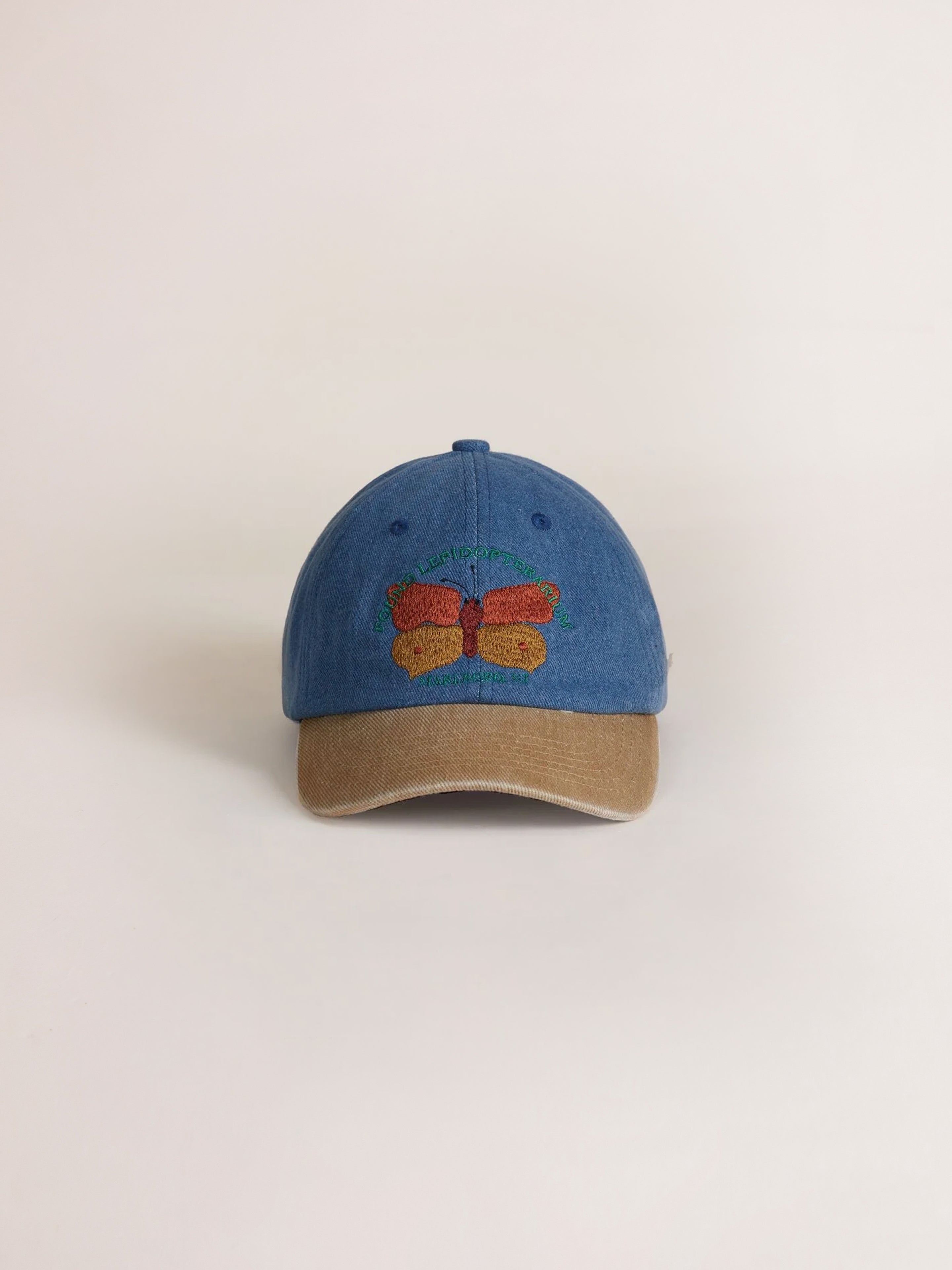 The Found Butterfly House Denim Cap features a blue baseball cap with a tan corduroy brim and intricate butterfly embroidery in multicolored threads on the front.