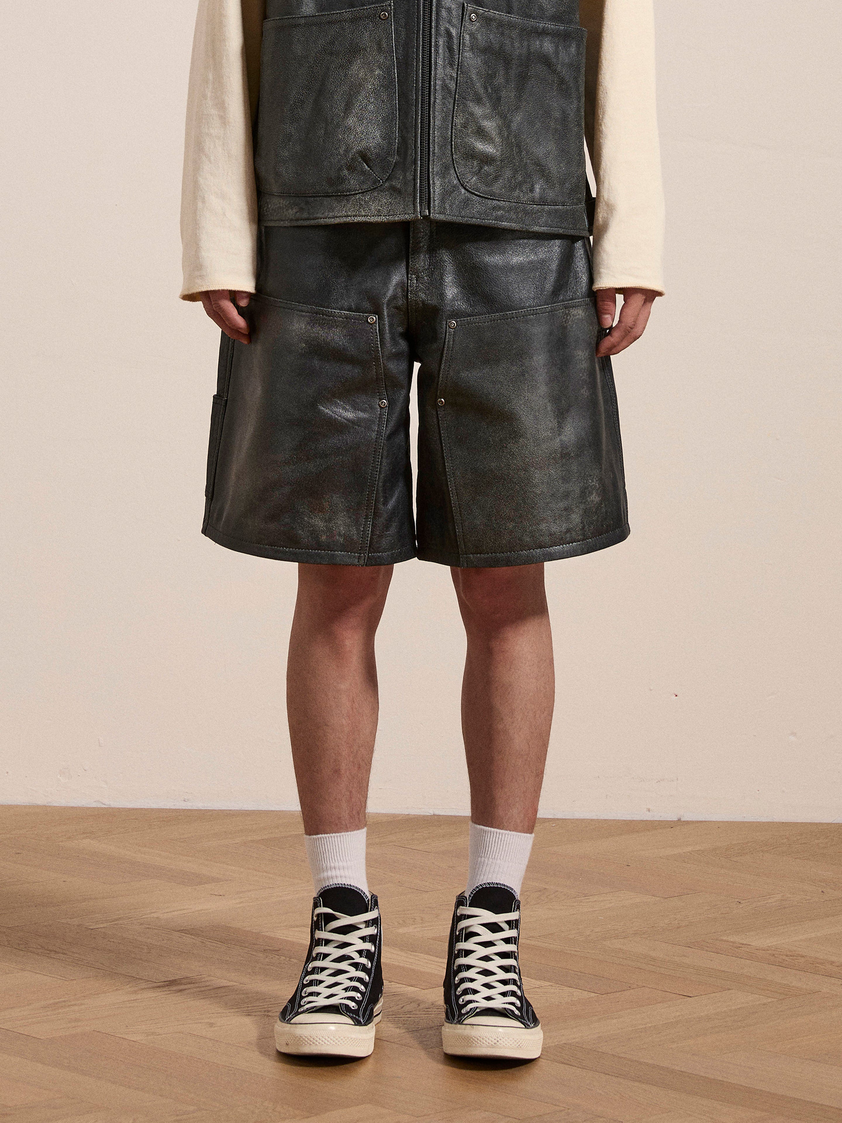 A person wearing a black leather vest and Found's Distressed Genuine Leather Carpenter Shorts in a double-knee silhouette stands on a wooden floor. They are also sporting white socks and black high-top sneakers. The background is plain.