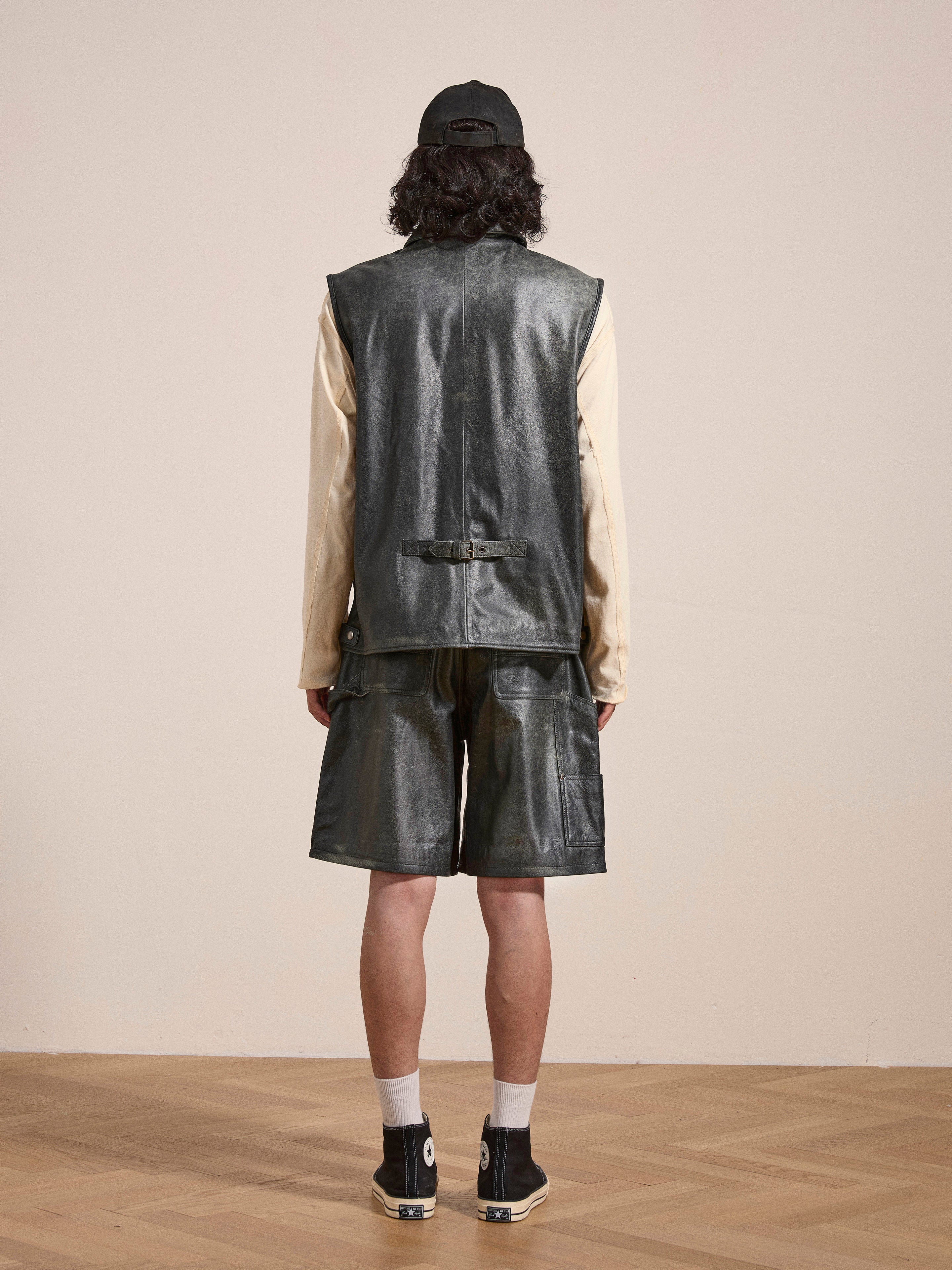 A person with shoulder-length hair is standing with their back to the camera, wearing a black cap, a sleeveless leather top, matching Distressed Genuine Leather Carpenter Shorts from Found, white socks, and black sneakers.