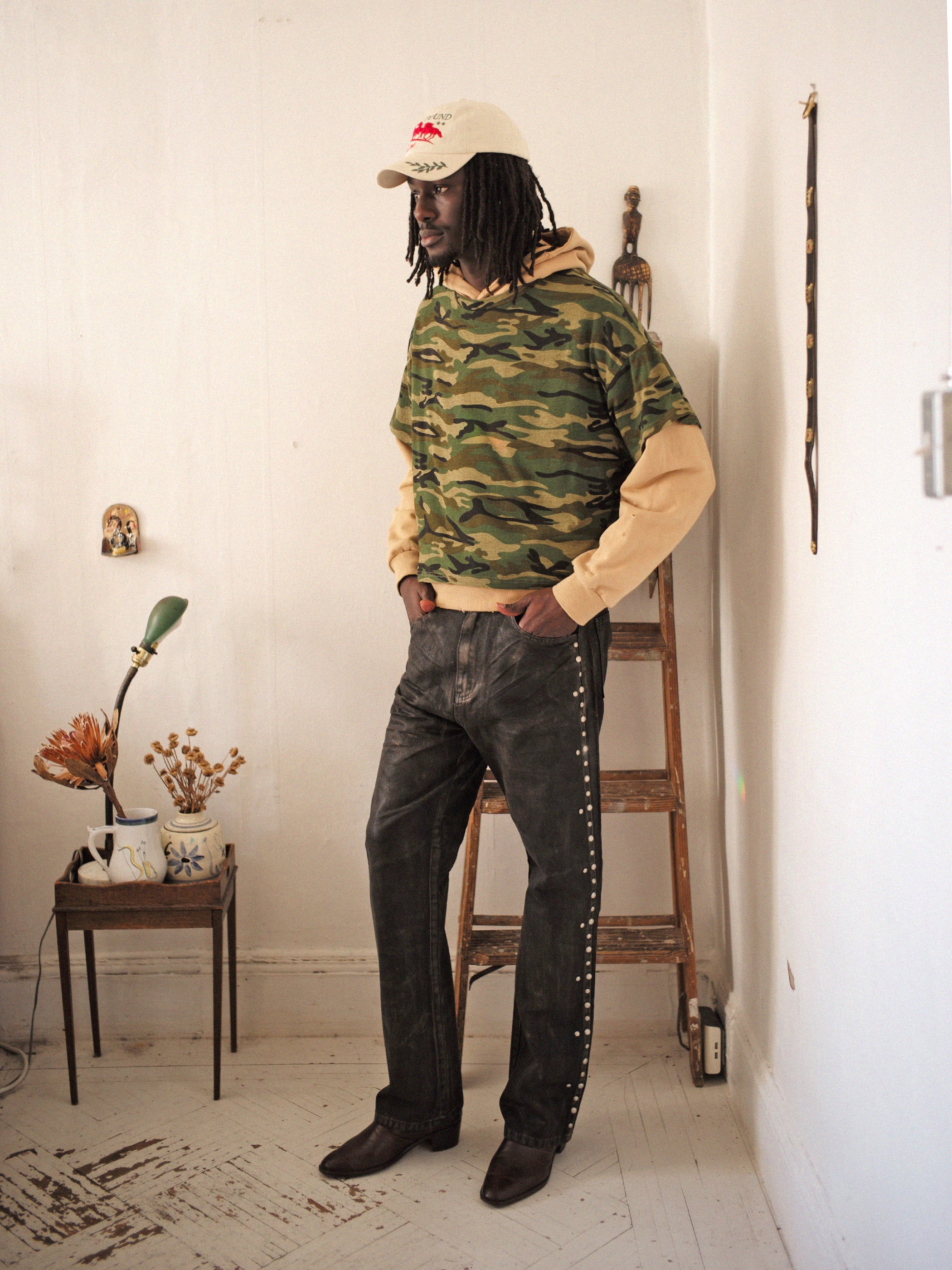 A person wears a vintage look, FOUND Double Layer Hoodie crafted from French Terry Cotton, jeans, and a beige cap while standing indoors near a wooden ladder and a decoratively adorned small table.