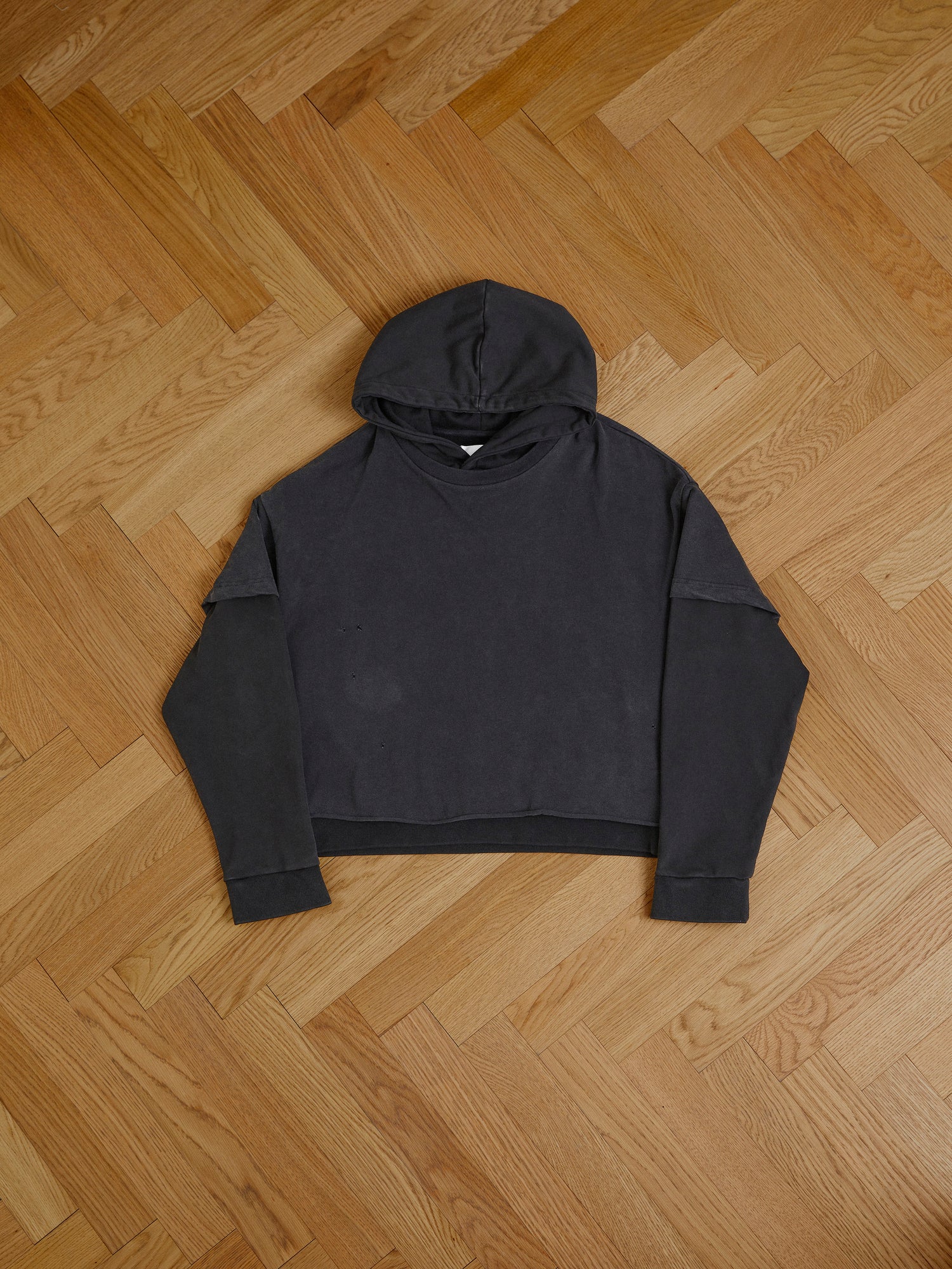 A black Found Double Layer Hoodie laid flat on a herringbone-patterned wooden floor.