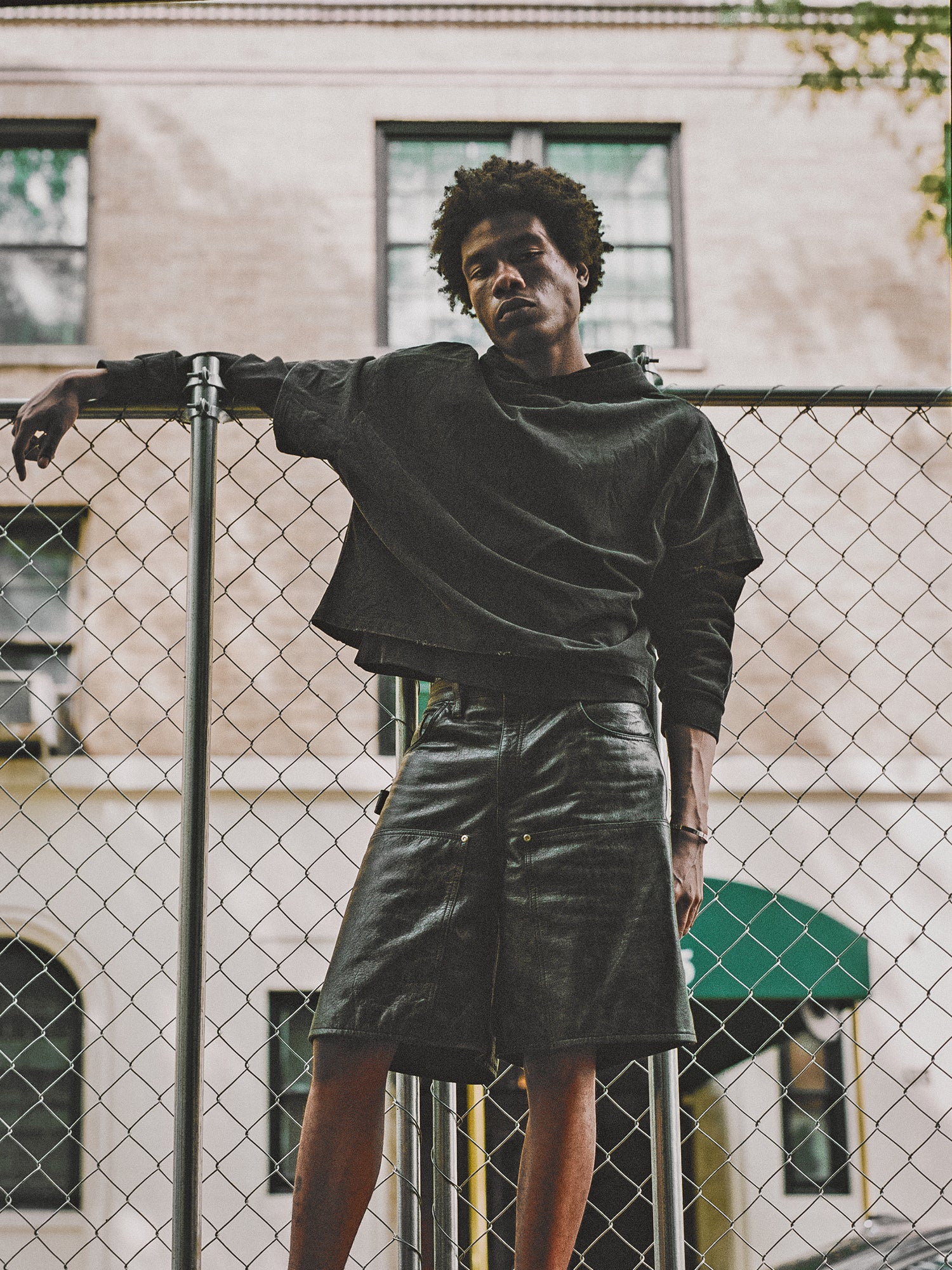 A person with short, curly hair leans against a chain-link fence, wearing a black Double Layer Hoodie by Found made of French terry cotton and black shorts. A building is visible in the background.