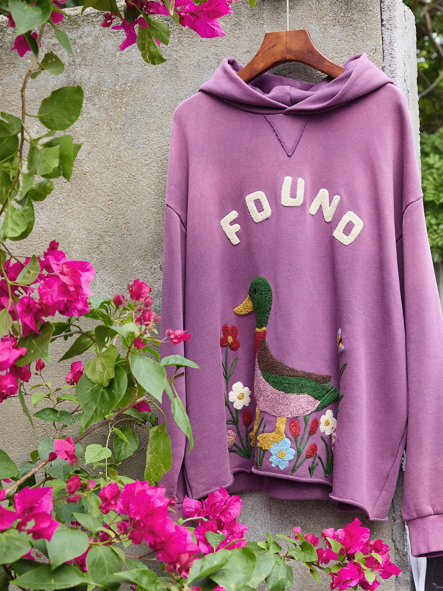 The Embroidered Wildlife Hoodie by FOUND, featuring duck and flower embroidery in mauve, hangs on a hanger near a wall and blooming pink bougainvillea.