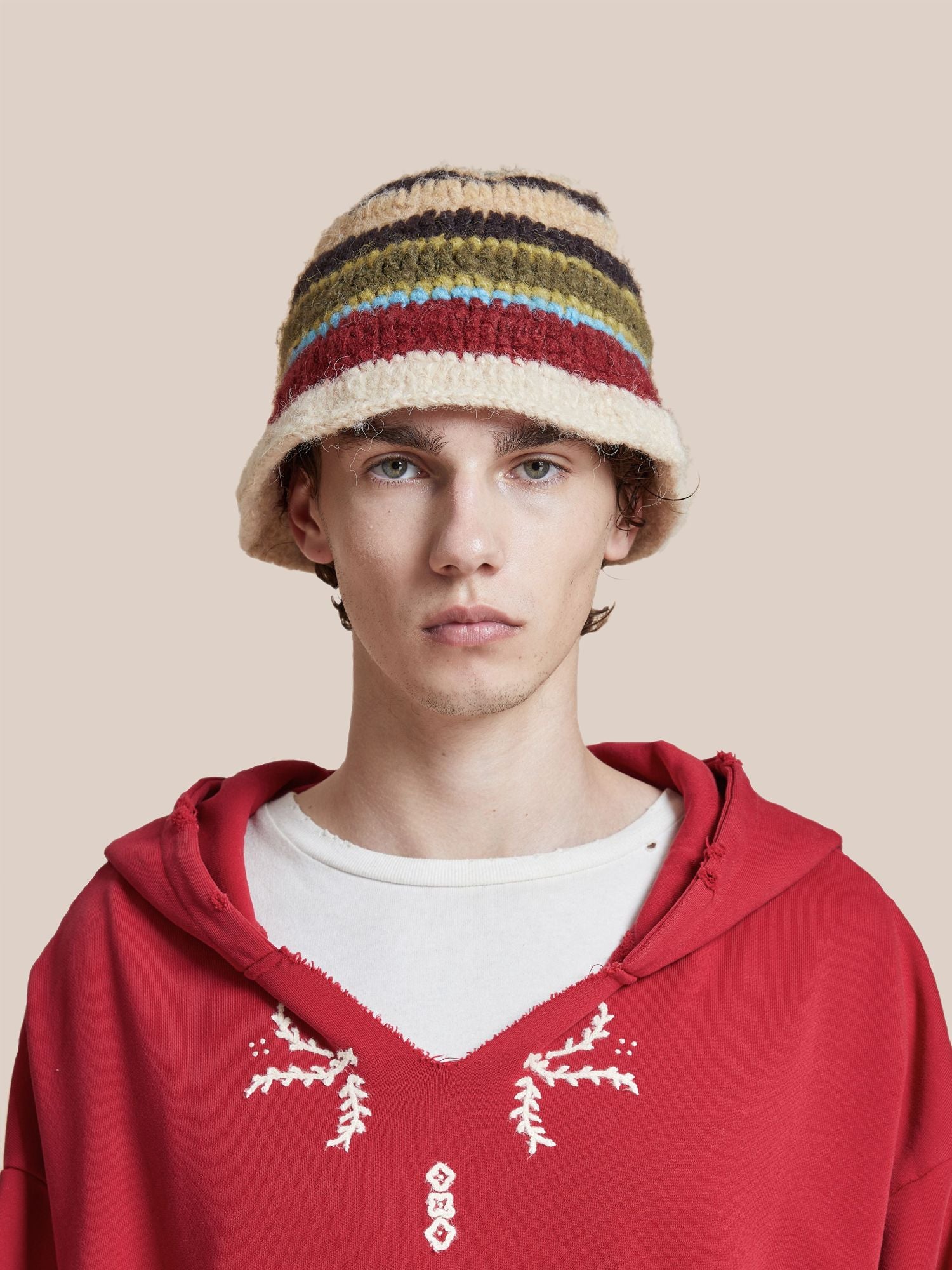 Young man in a red hoodie and a Found multi-color stripe knit beanie, staring at the camera against a light pink background.