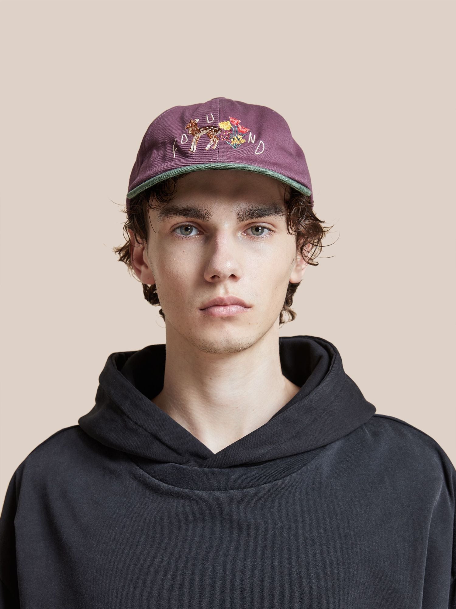 A young man wearing a maroon Found Flower Deer Cap and black hoodie, looking at the camera with a neutral expression on a light pink background.