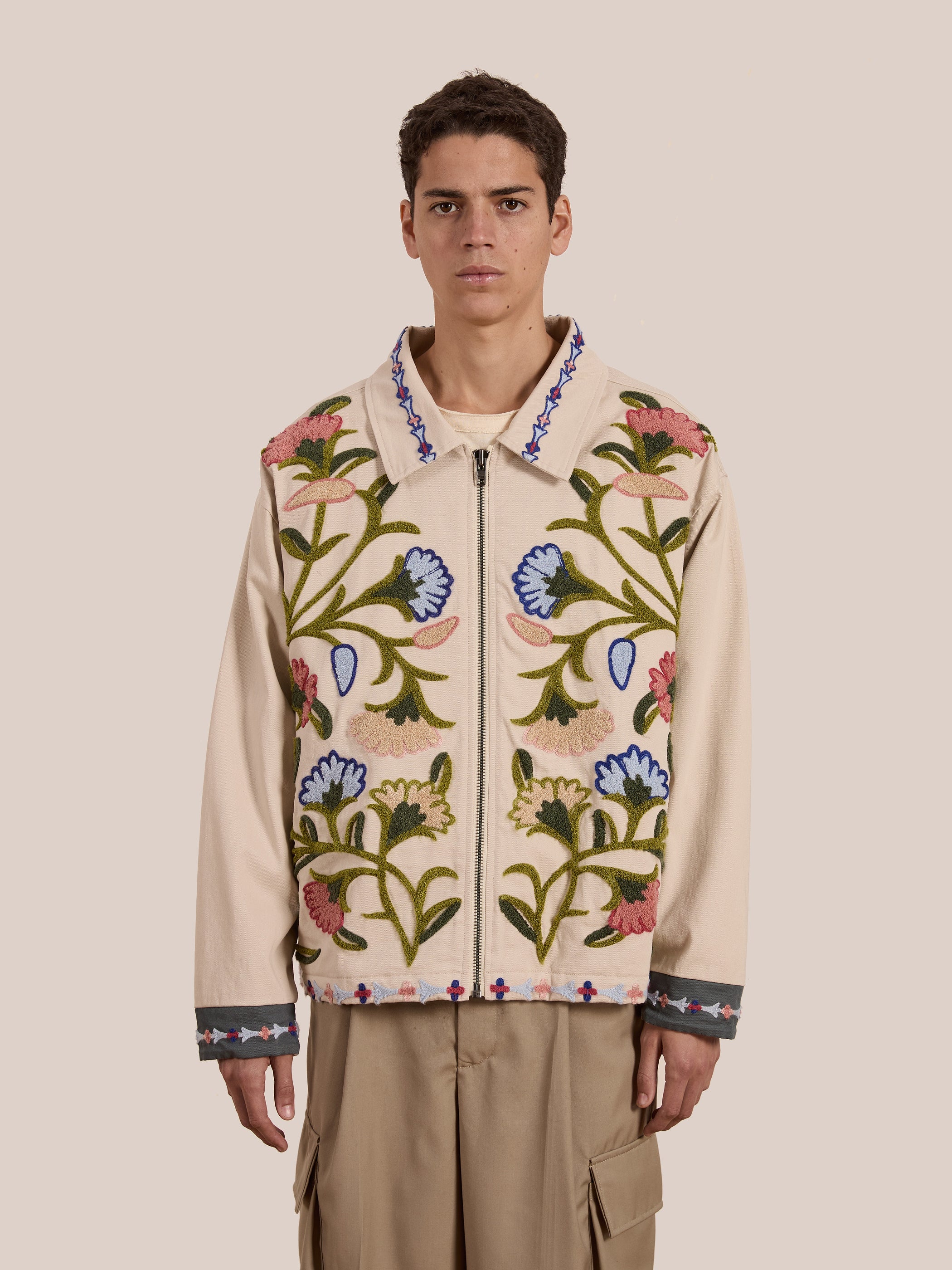 A person is standing against a plain background wearing FOUND's Floral Embroidered Work Jacket featuring vibrant chainstitch embroidery, paired with khaki pants.