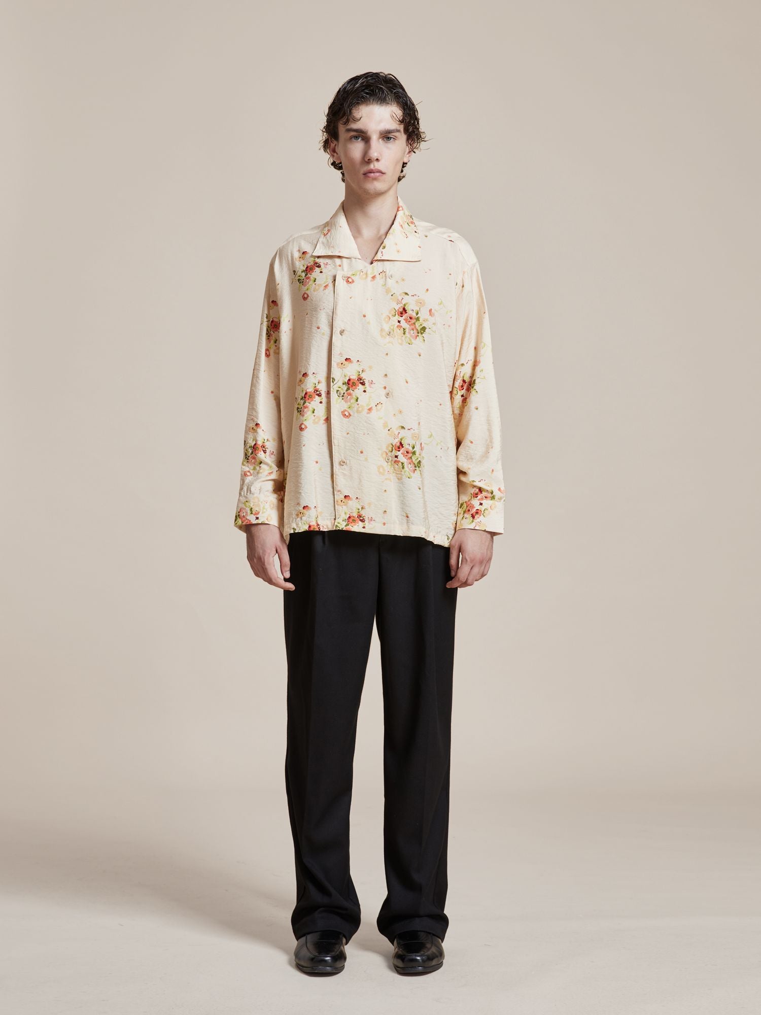 A person with short, wavy hair is standing and facing forward. They are wearing the Found Kanhati Garden Long Sleeve Camp Shirt adorned with delicate Phulkari motifs and black pants. The background is plain beige, highlighting the intricate designs inspired by Punjab, Pakistan.