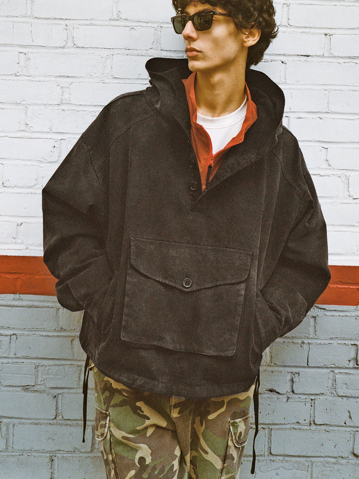 Clad in a FOUND Washed Twill Pocket Anorak Jacket with a unisex relaxed fit, camouflage pants, and sunglasses, someone stands confidently against a brick wall.