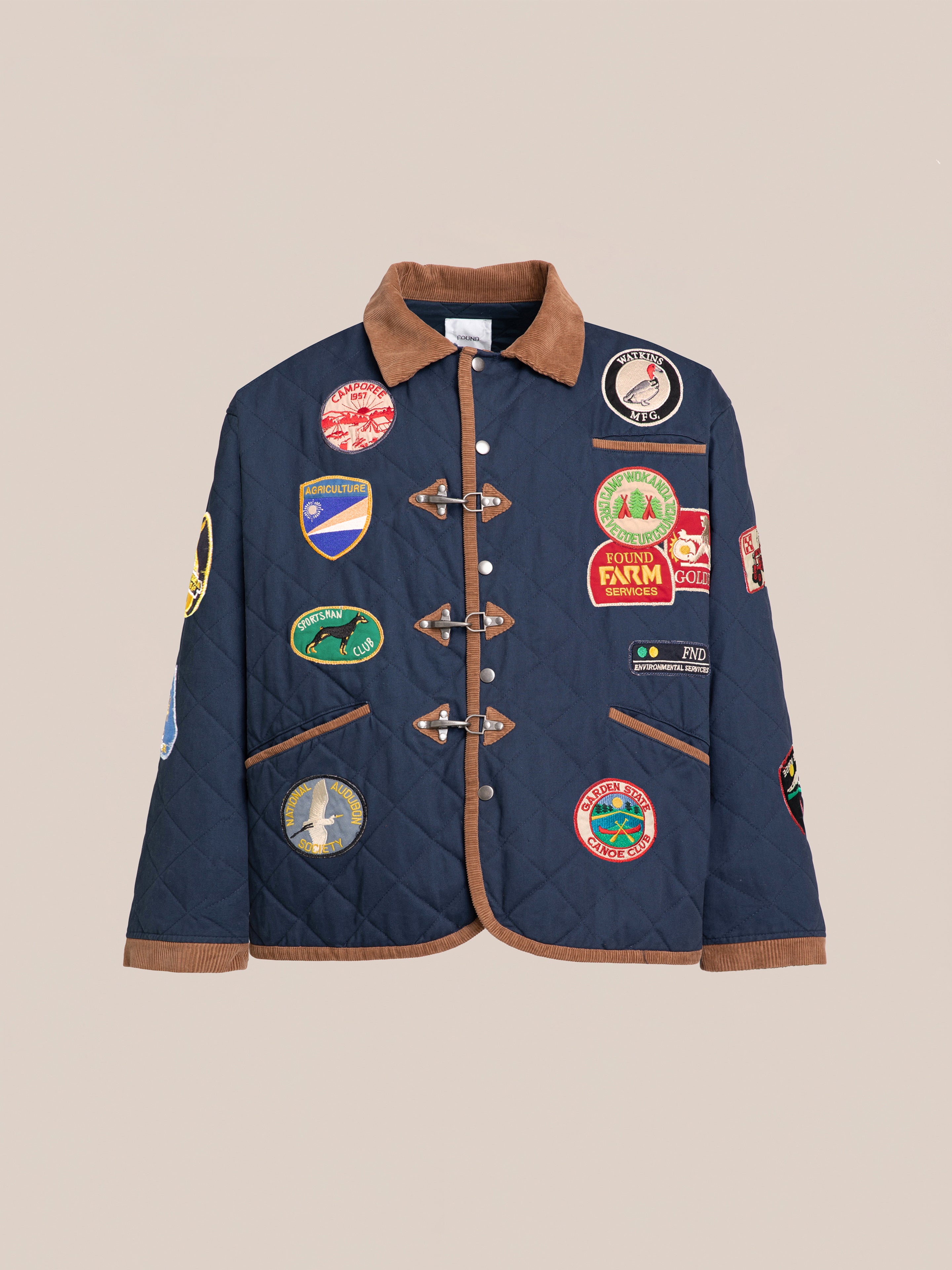 The Farmstead Quilt Patch Jacket by FOUND is a navy blue quilted jacket adorned with a brown collar and cuffs. It features multiple colorful patches reminiscent of vintage hunting competition jackets and has toggle closures on the front.