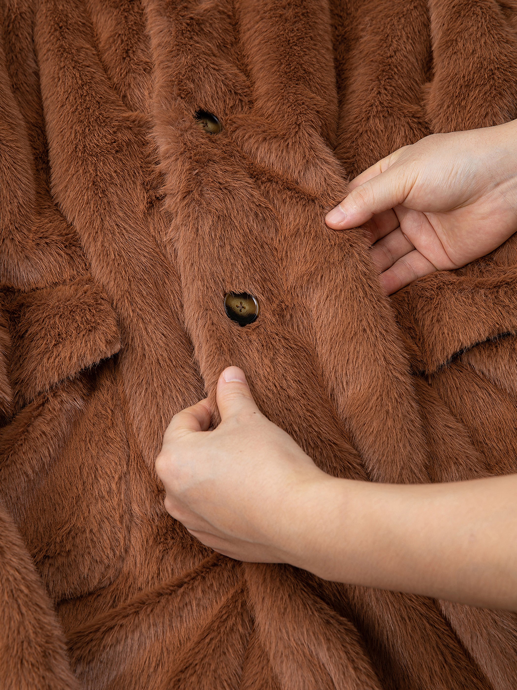 Hands adjusting buttons on the FOUND Long Faux Fur Coat in brown, revealing a glimpse of its luxurious satin lining.