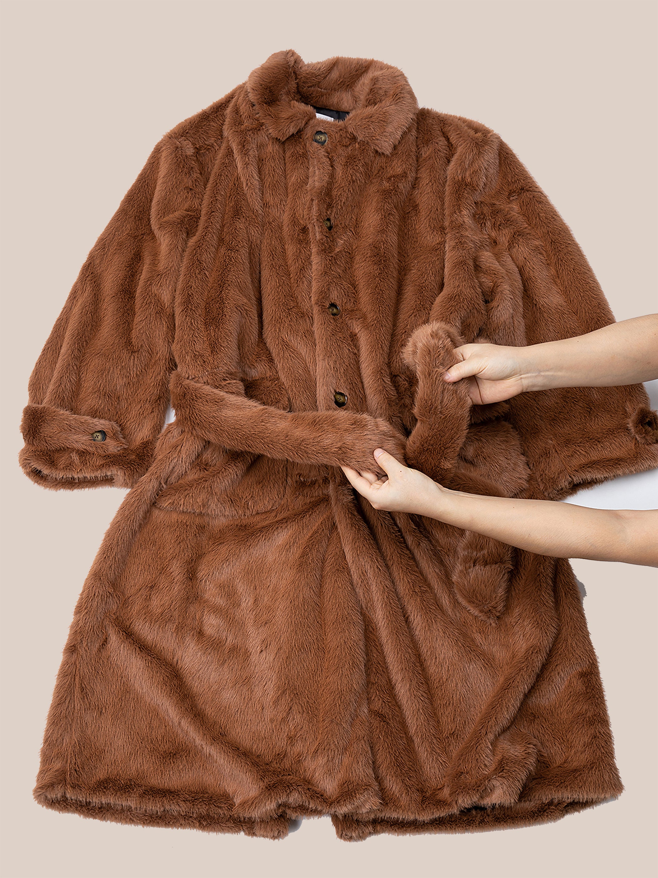 Two hands hold the belt of the FOUND Long Faux Fur Coat, a double-breasted design in brown with buttons and long sleeves, laid out on a neutral background.