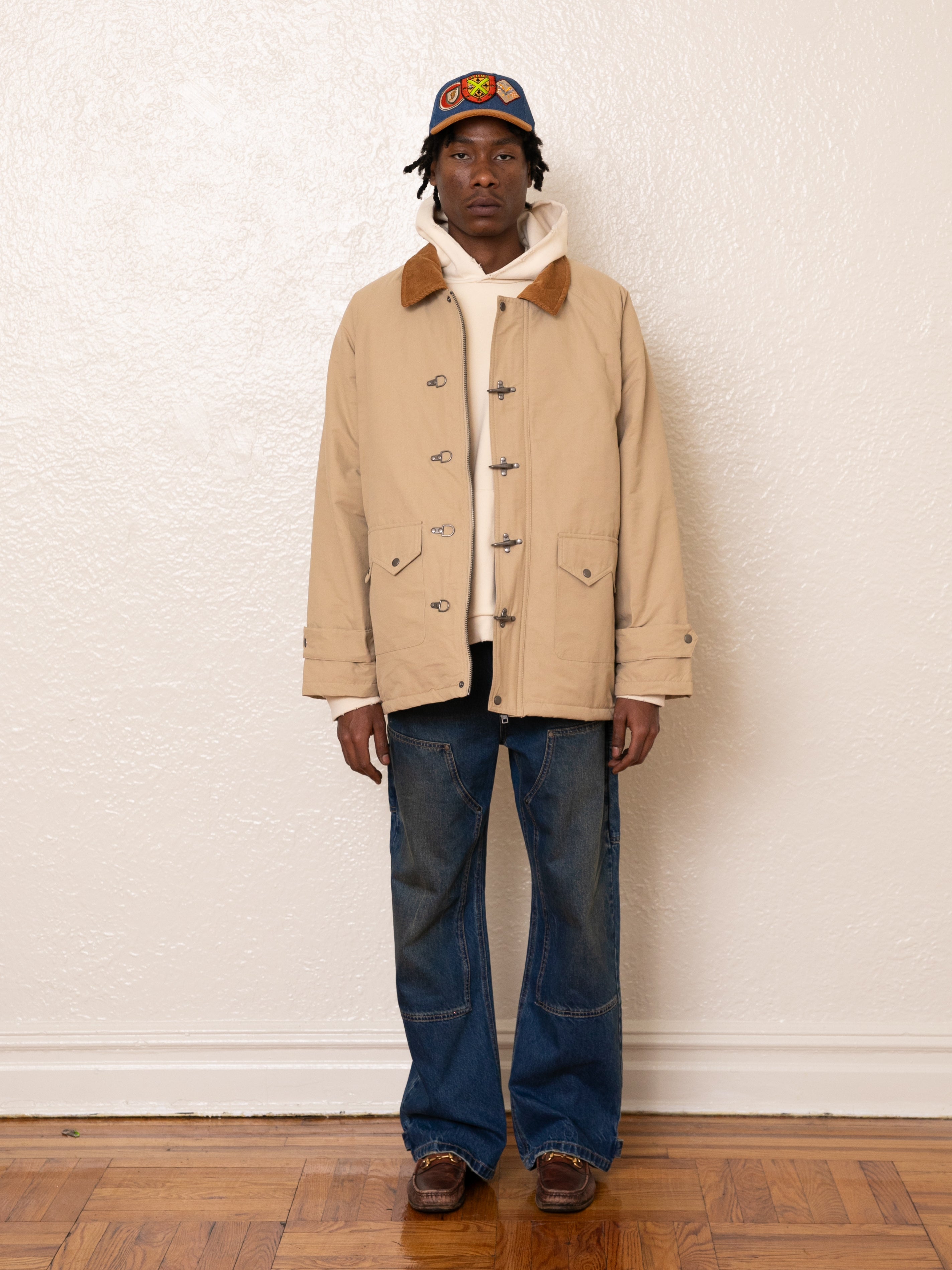 A person is indoors wearing FOUND's Fisherman Jacket with a corduroy collar, paired with a white hoodie, blue jeans, and a colorful cap. The wooden floor contrasts with the plain white wall.