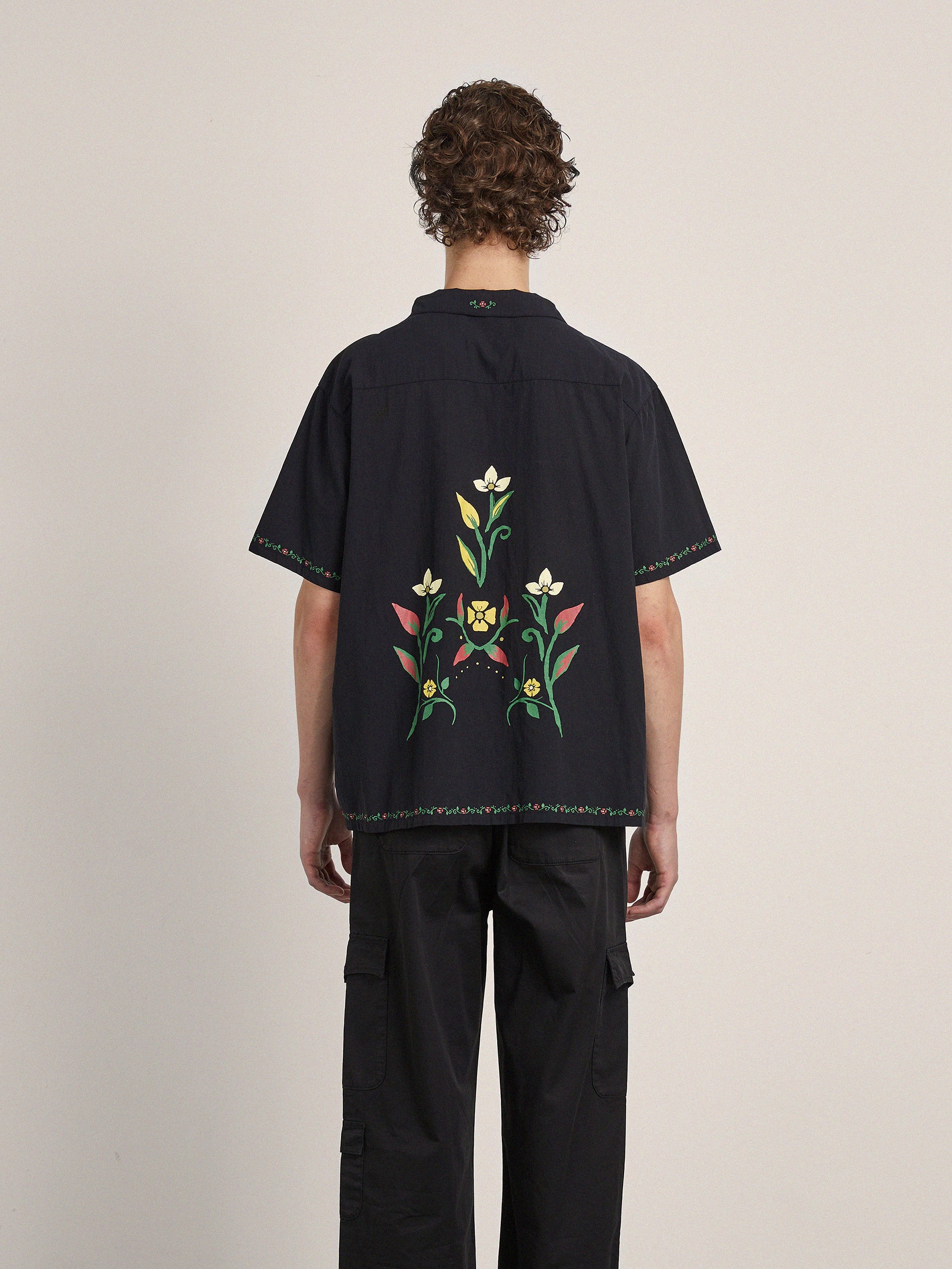 A person with curly hair is seen from the back wearing a black Floral Camp Shirt by Profound with intricate embroidery, made from 100% cotton fabric, paired with relaxed fit black cargo pants.