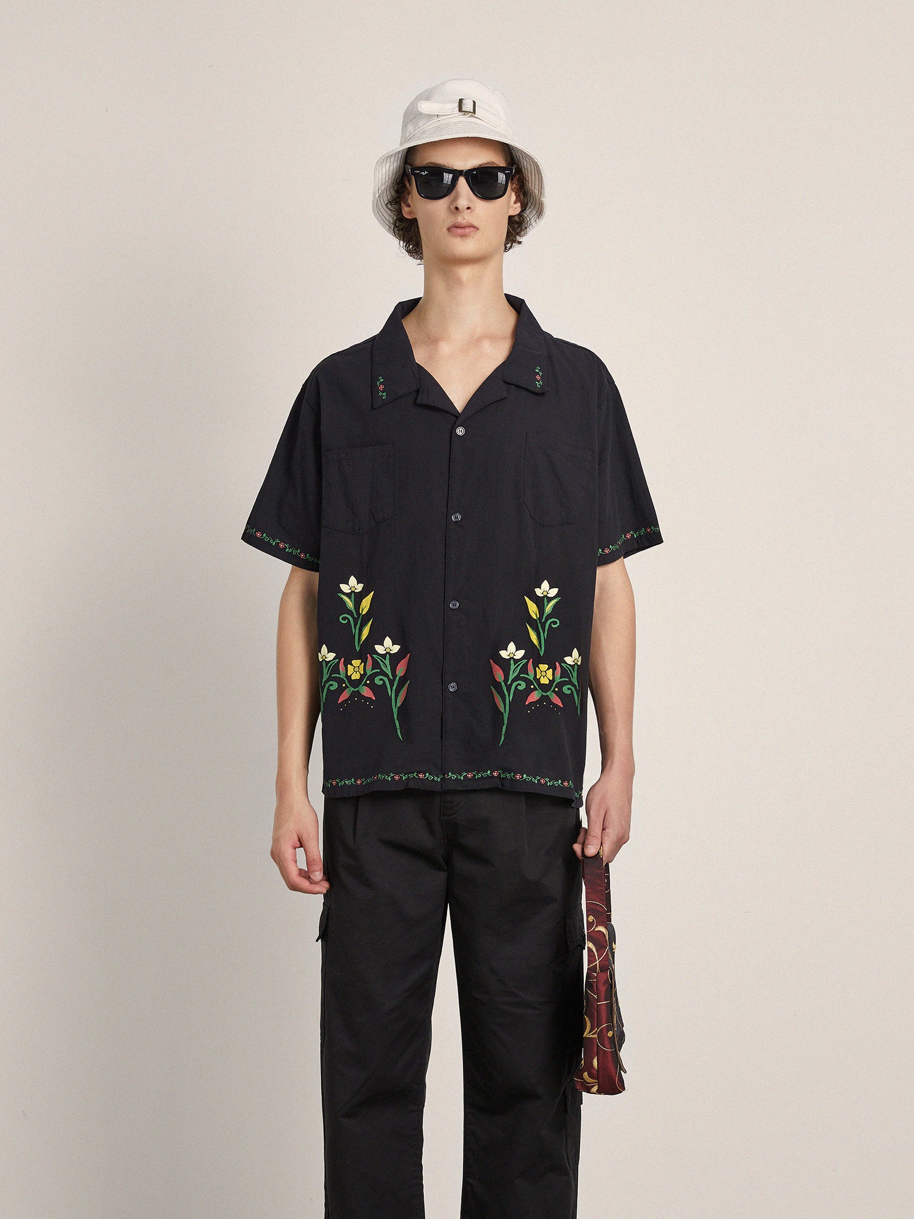 A person wearing a black Floral Camp Shirt from Profound made from 100% cotton fabric with floral embroidery, relaxed fit black pants, a white bucket hat, and sunglasses.