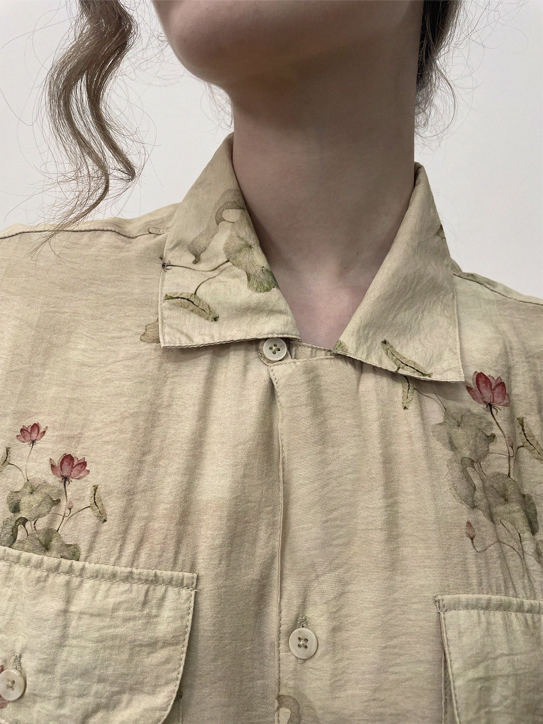 Close-up of a person wearing a beige Found Lotus LS Camp Shirt with floral embroidery and dual chest pockets. The hair is styled in loose curls, and the photo captures the details of the shirt's front button closure against a plain background.