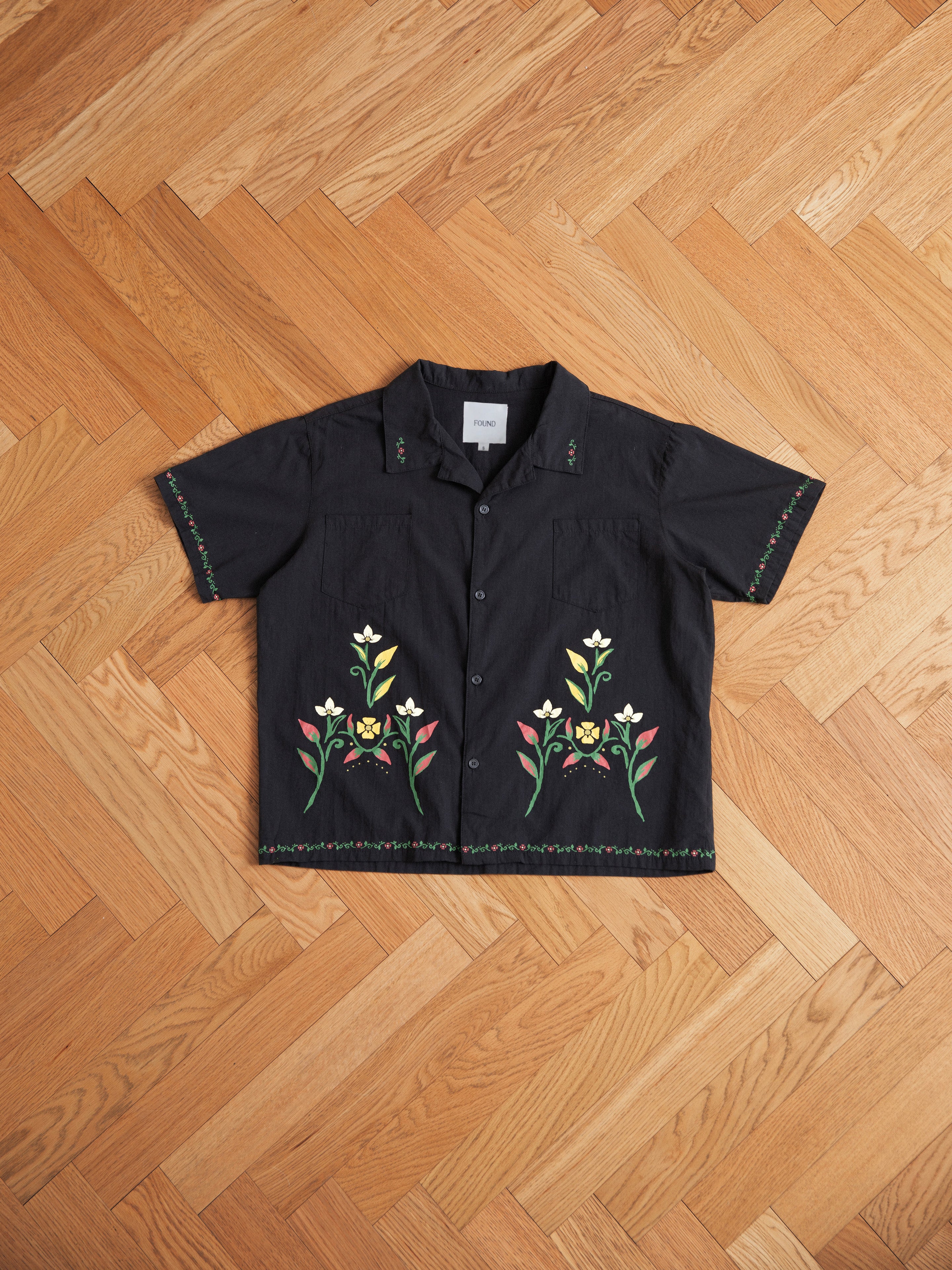 A black Floral Camp Shirt from Profound with short sleeves and floral embroidery on the front and sleeves, crafted from 100% cotton. Its relaxed fit makes it perfect for any casual occasion, displayed beautifully on a wooden herringbone floor.