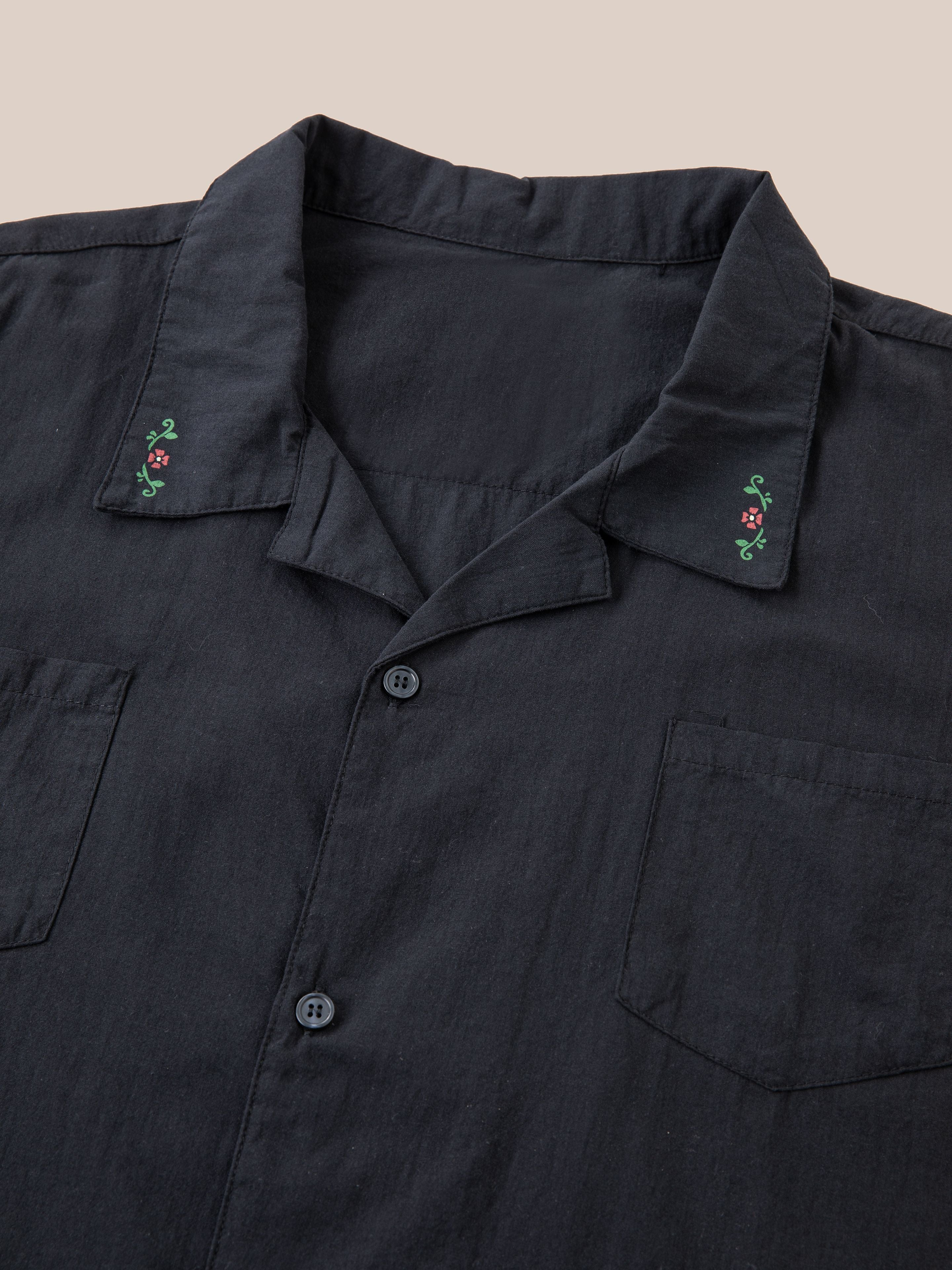 A black Profound Floral Camp Shirt with a pointed collar featuring small floral embroidery on each side. This relaxed fit button-up shirt, made from 100% cotton, has two chest pockets and is laid flat on a neutral background.