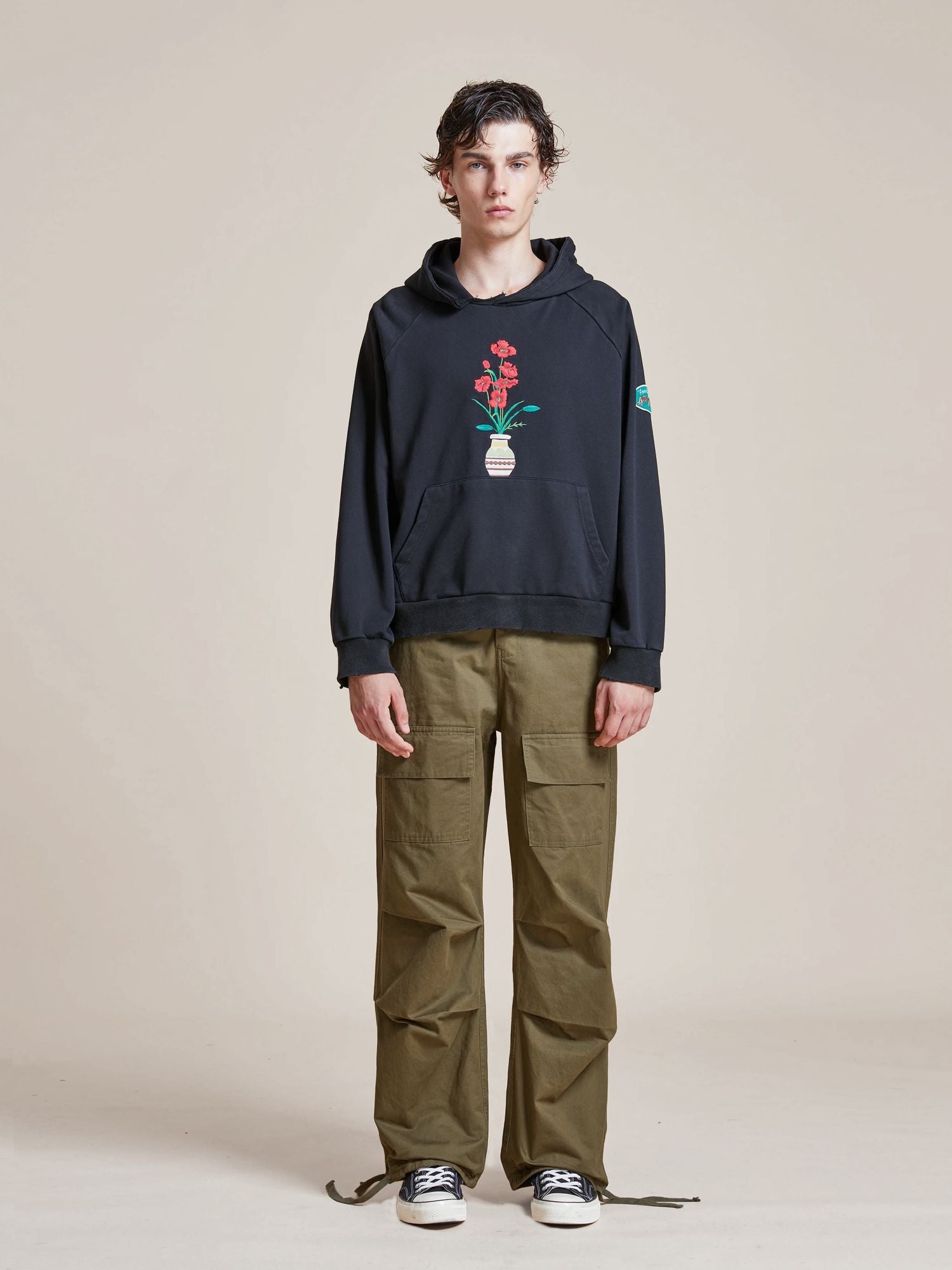 A young person stands against a neutral background, wearing a black Found Flowers Vase Hoodie with a flower pot design, olive green cargo pants, and black-and-white sneakers, giving off a subtle vintage feel.