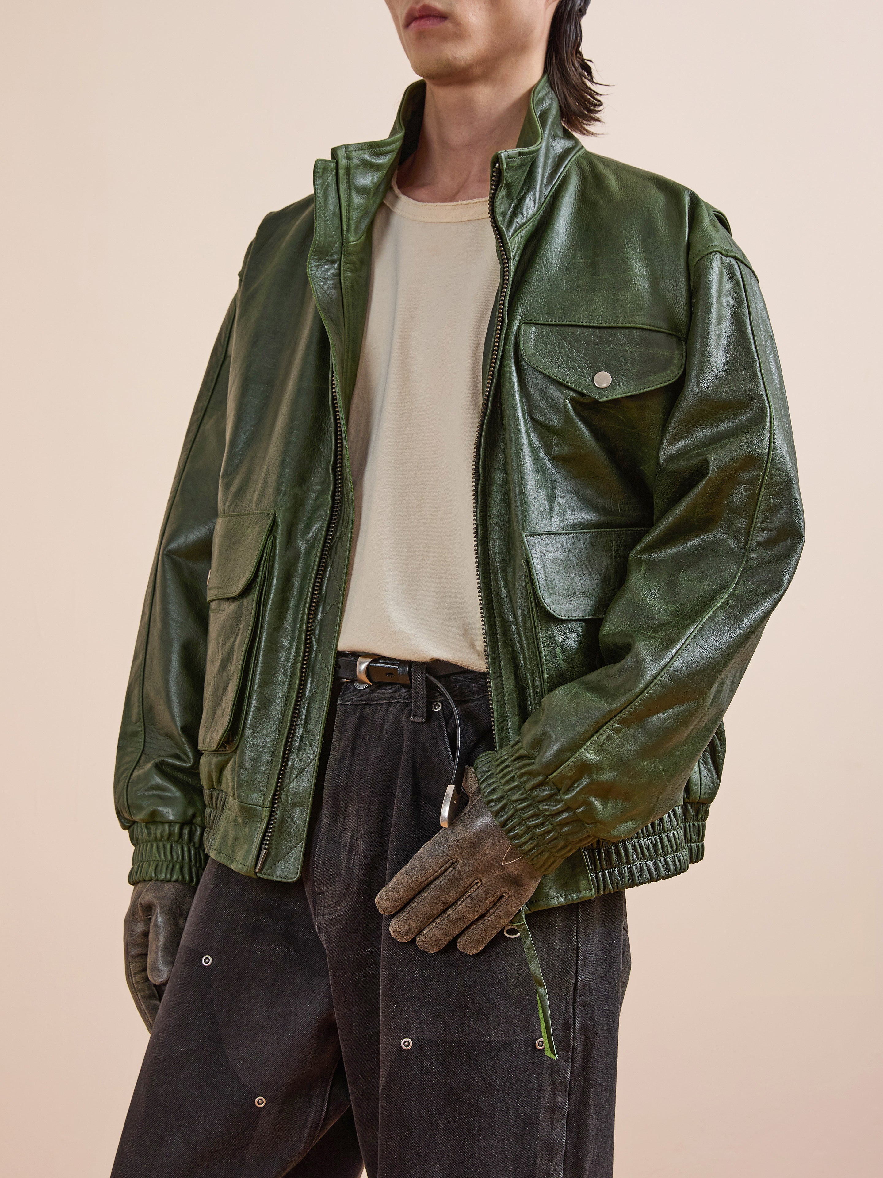 A person wearing the FOUND Forest Leather Bomber Jacket in green genuine leather over a white t-shirt, black pants, and gloves stands against a plain beige background.