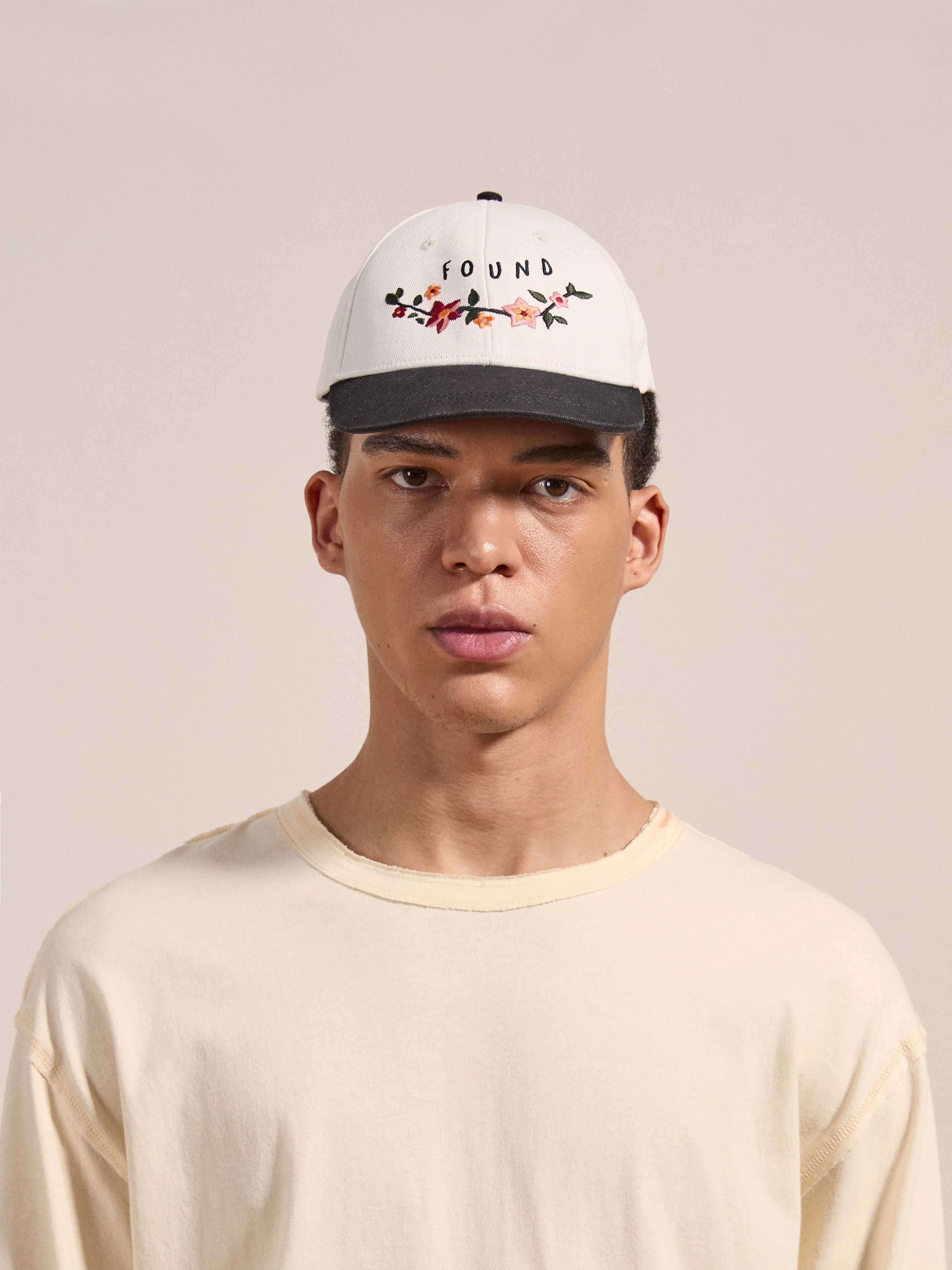 A person wearing a plain beige shirt and the Greenlands Contrast Cap, featuring the brand name "FOUND" along with floral embroidery, standing against a neutral background.