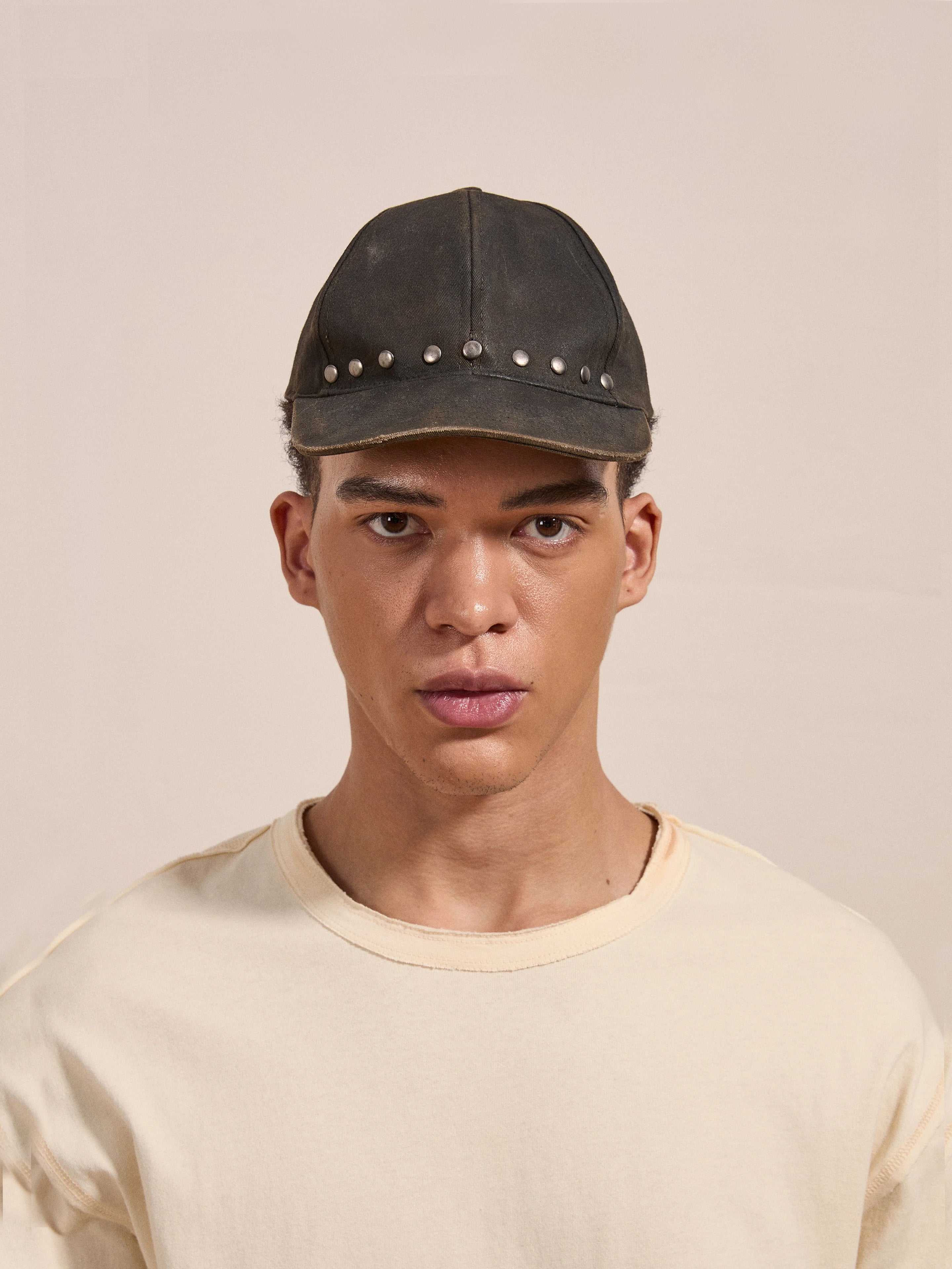 Individual wearing the "FOUND" Waxed Charcoal Studded Cap and a beige t-shirt, staring directly into the camera against a plain background.