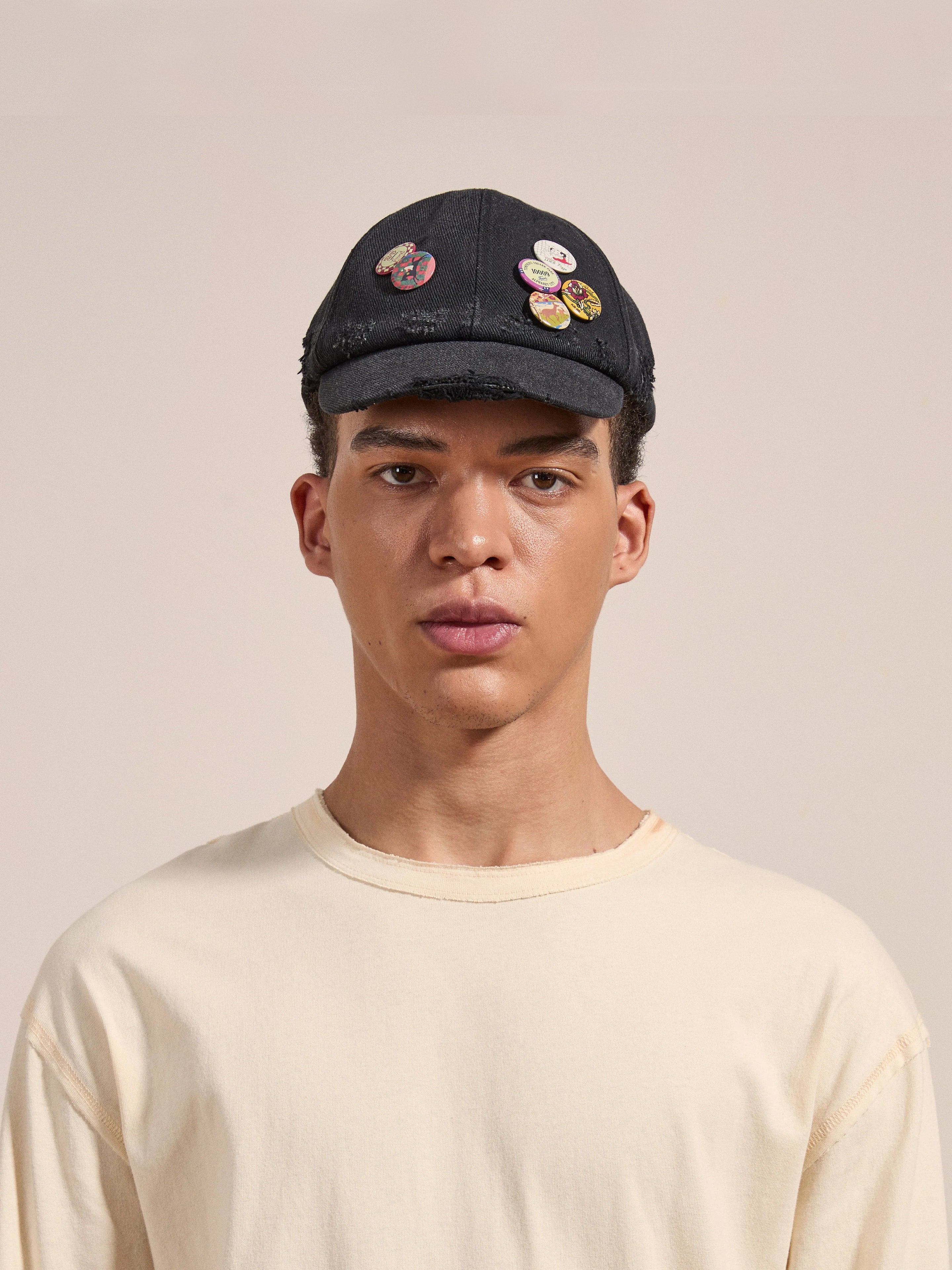 A person wearing a cream-colored shirt and FOUND's Distressed Pin Cap adorned with several colorful pins, standing against a plain background.