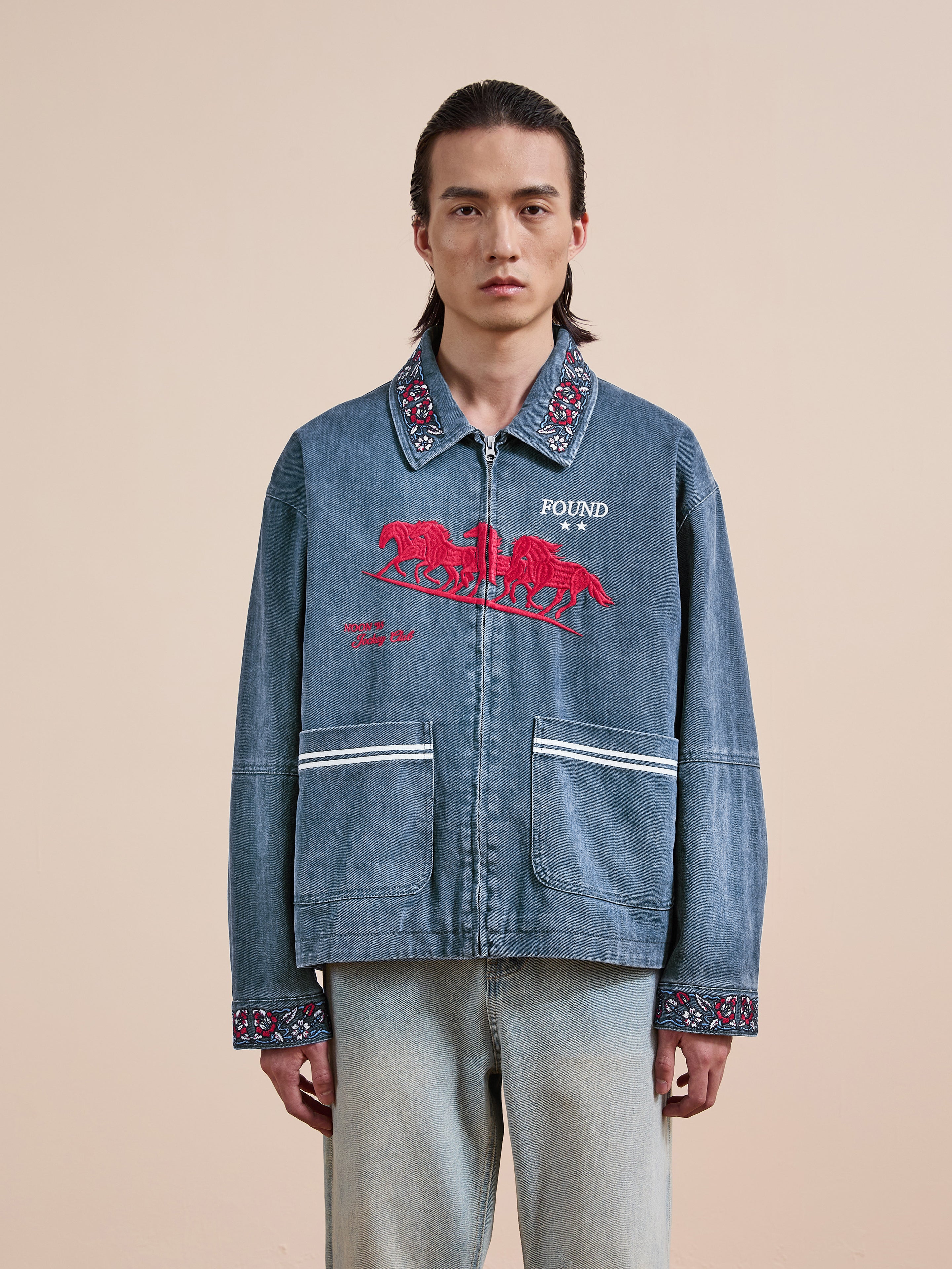 Wearing the Found Jockey Club Embroidered Jacket in blue denim, featuring racing horses embroidery and decorative trim, paired with light-wash jeans, the person stands against a plain beige background.