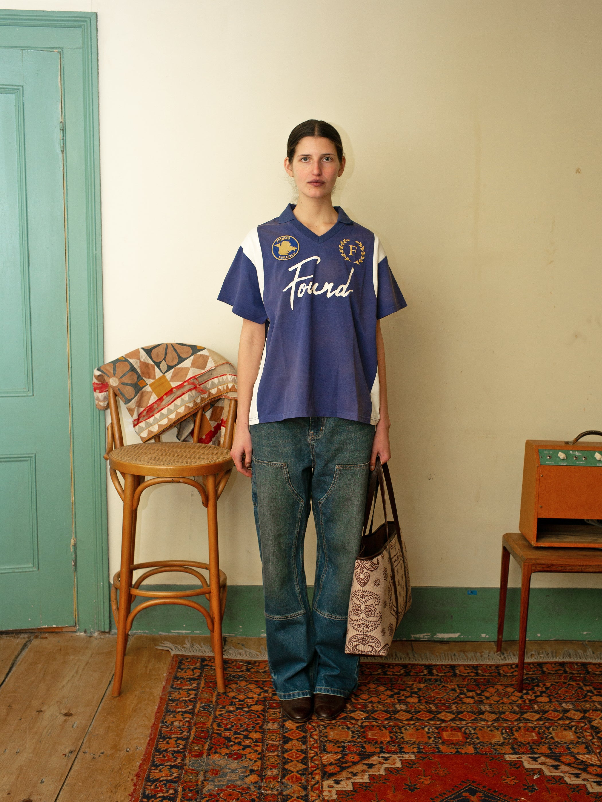 A person stands in a room wearing the FOUND Premier League Jersey, paired with jeans and holding a patterned bag. Beside them is a chair draped with cloth and a small cabinet, adding to the eclectic vibe.