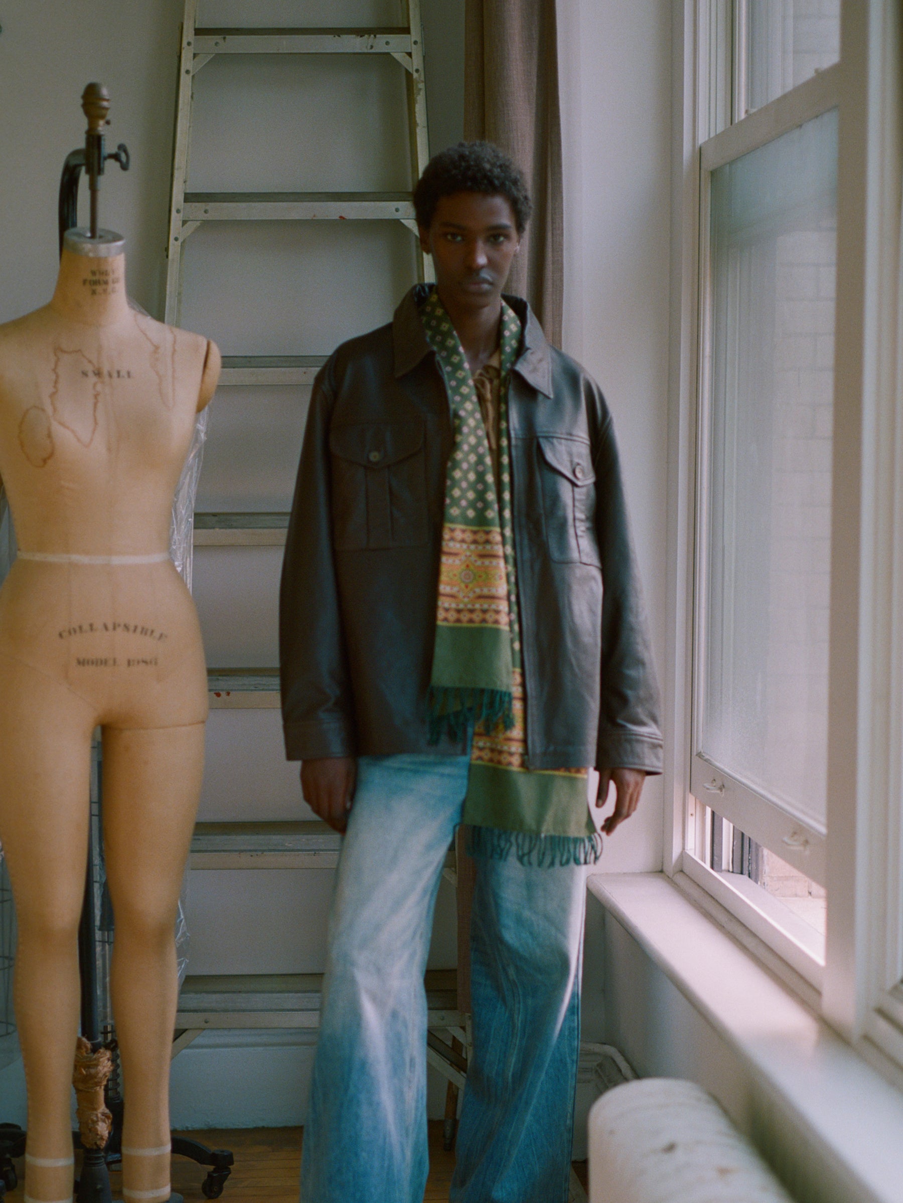 A mannequin adorned with Indo-Aryan prints stands beside a man, who is wearing a Found Greener Pastures Scarf with hand-tied tassels, in a room.