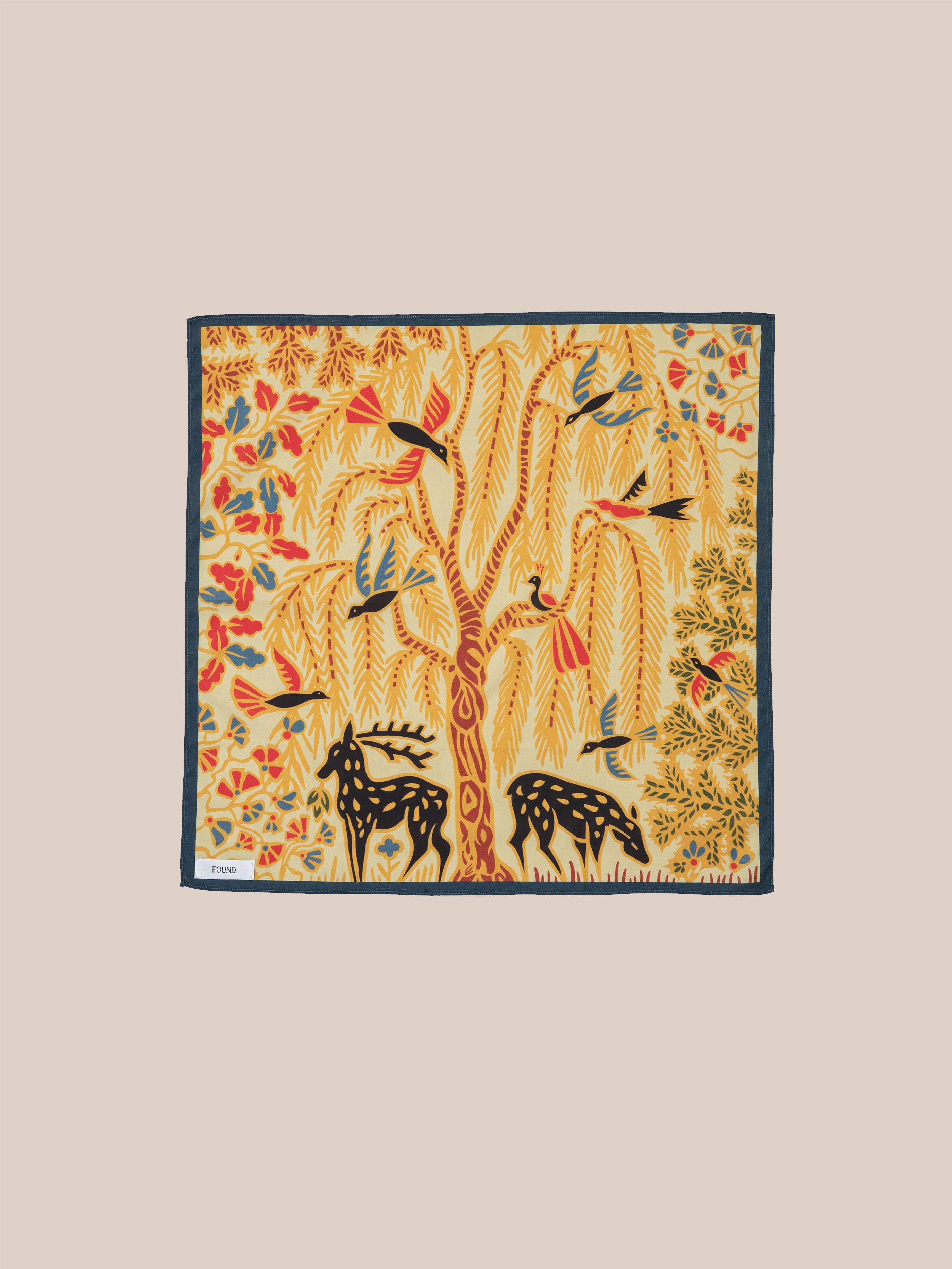 The Rainforest Bandana by Found features a vibrant nature-inspired design with a tree, birds, and two deer set against a yellow background with a dark border. Its intricate detailing is reminiscent of Phulkari artwork from the Punjab region.