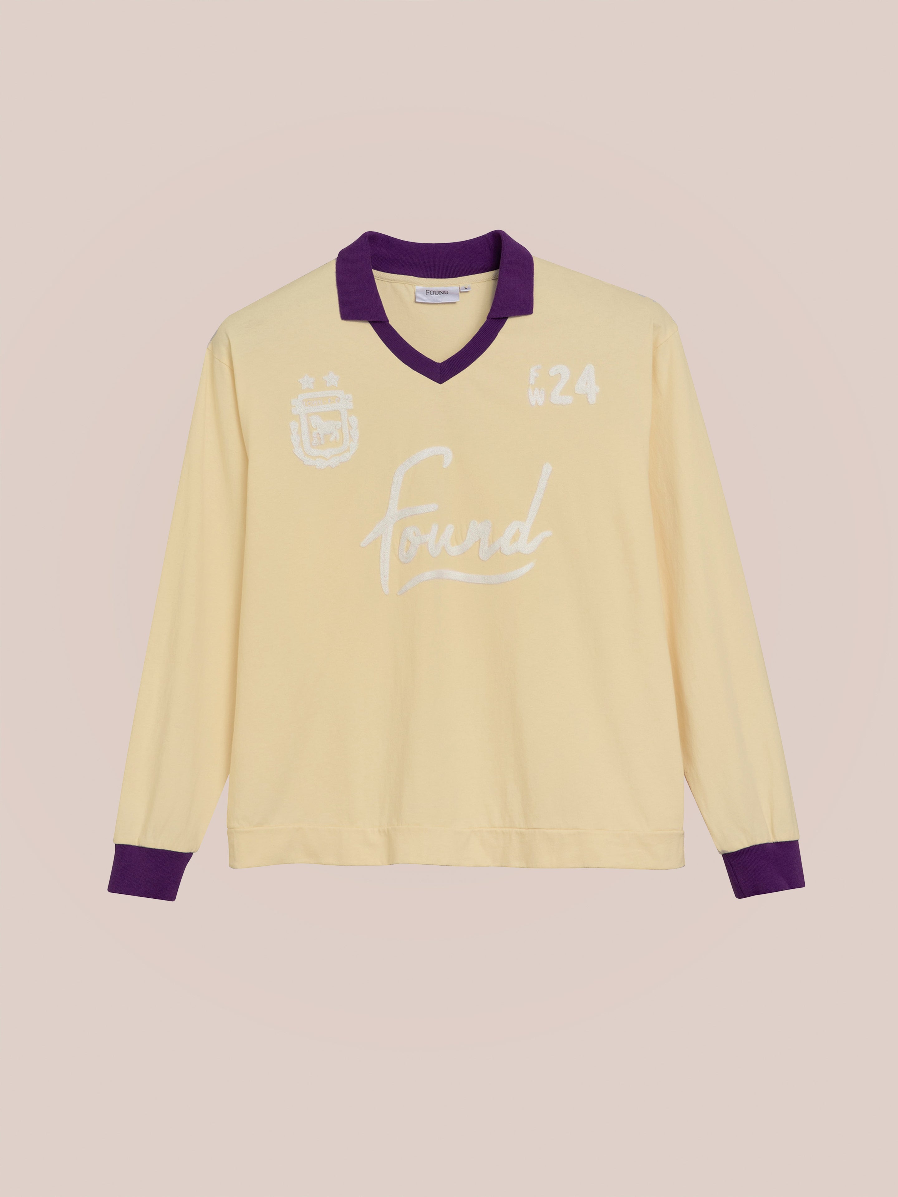 The Sahara Sun Jersey by FOUND is a long-sleeve yellow shirt with a purple collar and cuffs, featuring white text that reads "Found" and "F24" on the front. Reminiscent of vintage athletic uniforms, this piece combines classic style with modern flair.