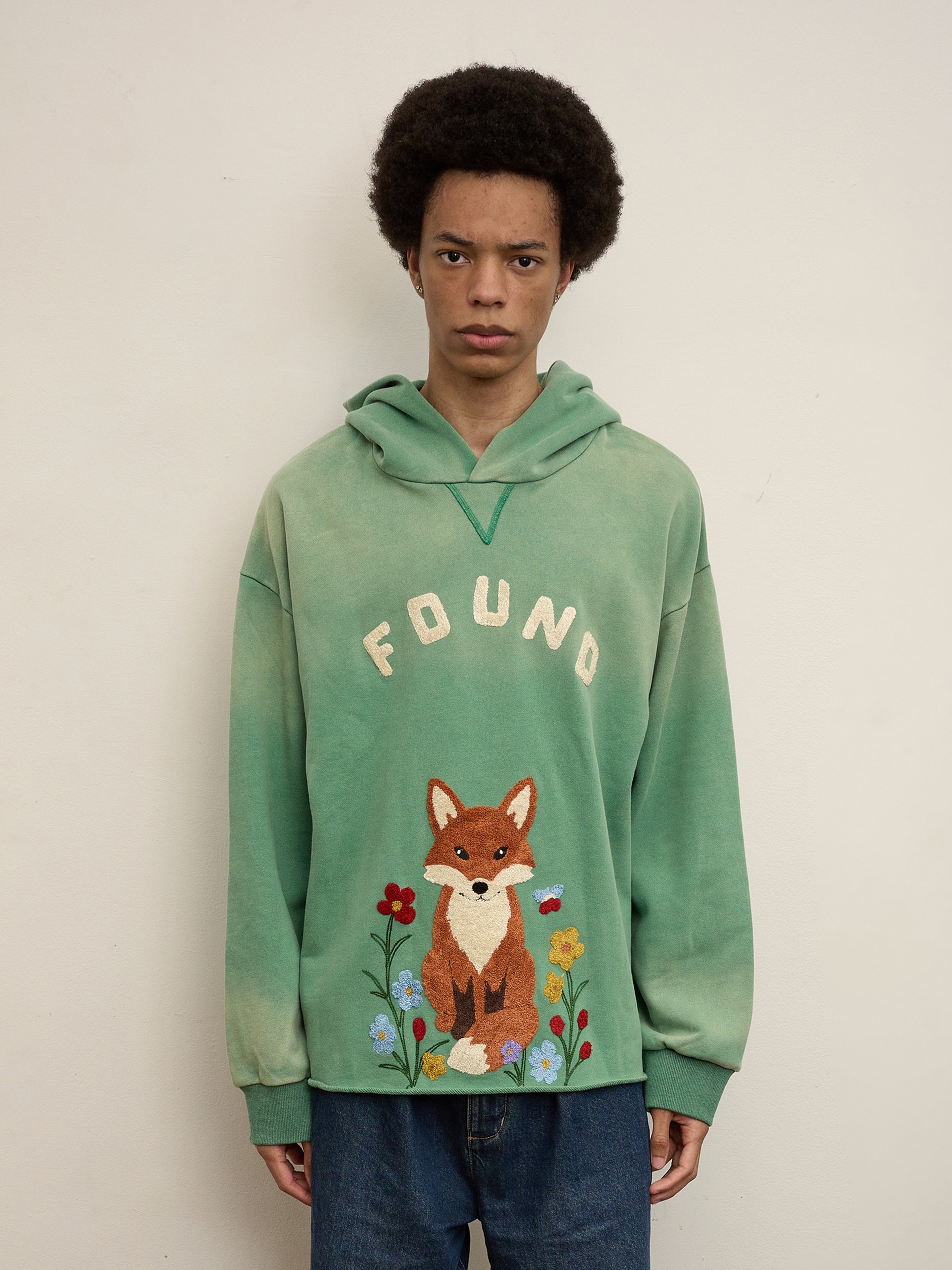 A person with an afro stands against a plain background, wearing a green FOUND Embroidered Wildlife Hoodie made from soft French terry cotton, featuring "FOUND" and embroidered fox and flowers on the front, paired with blue jeans.