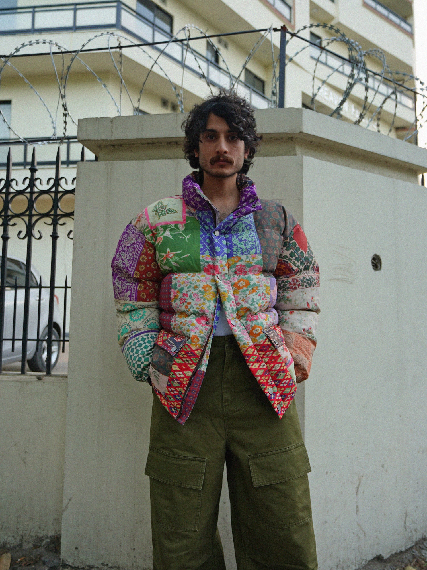 A man in a colorful Found Gardenia Tapestry Puffer Jacket standing next to a fence.