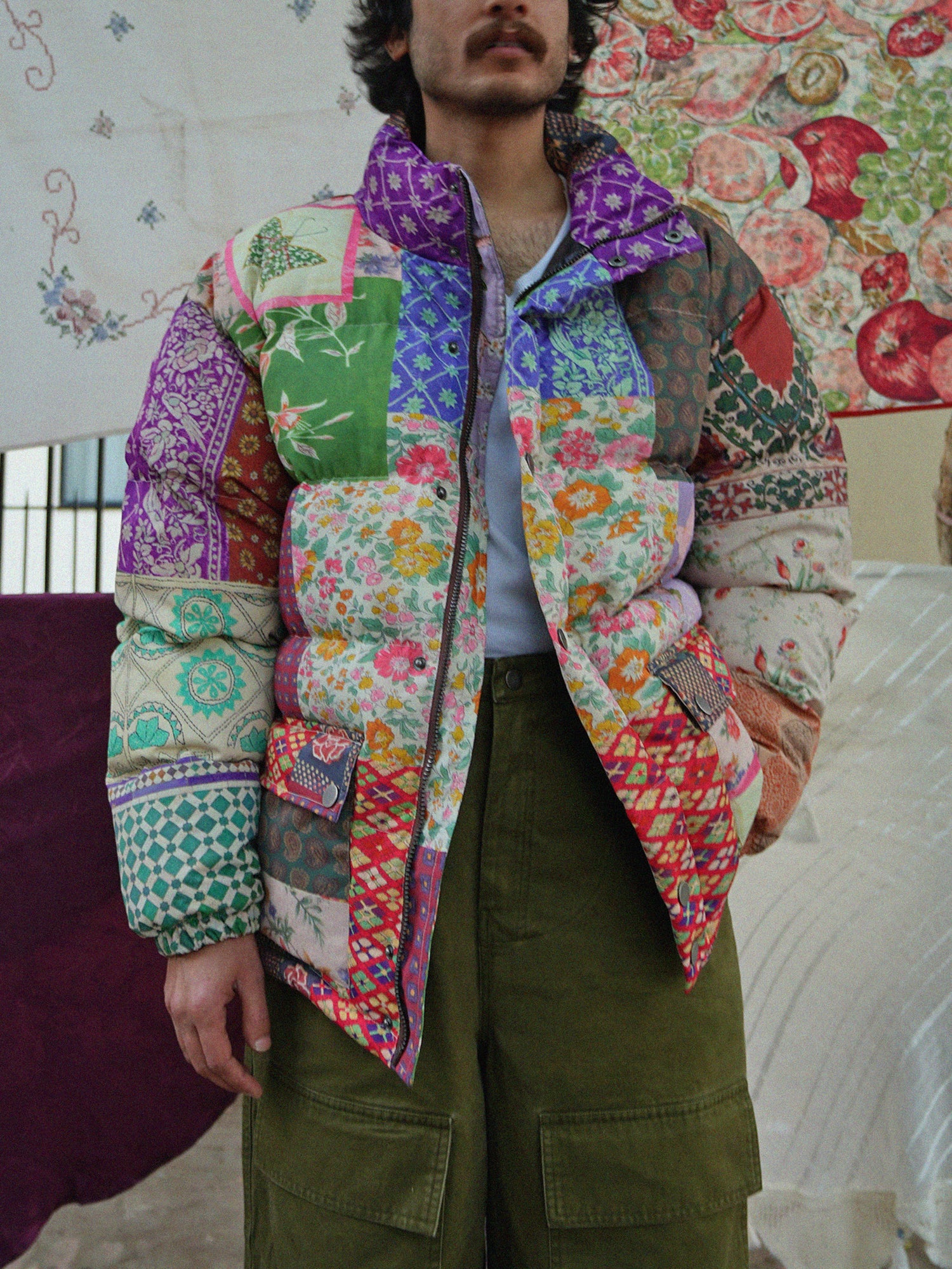A man standing in front of a pre-order item, the Found Gardenia Tapestry Puffer Jacket, showcasing unique South Asian prints on a quilt.