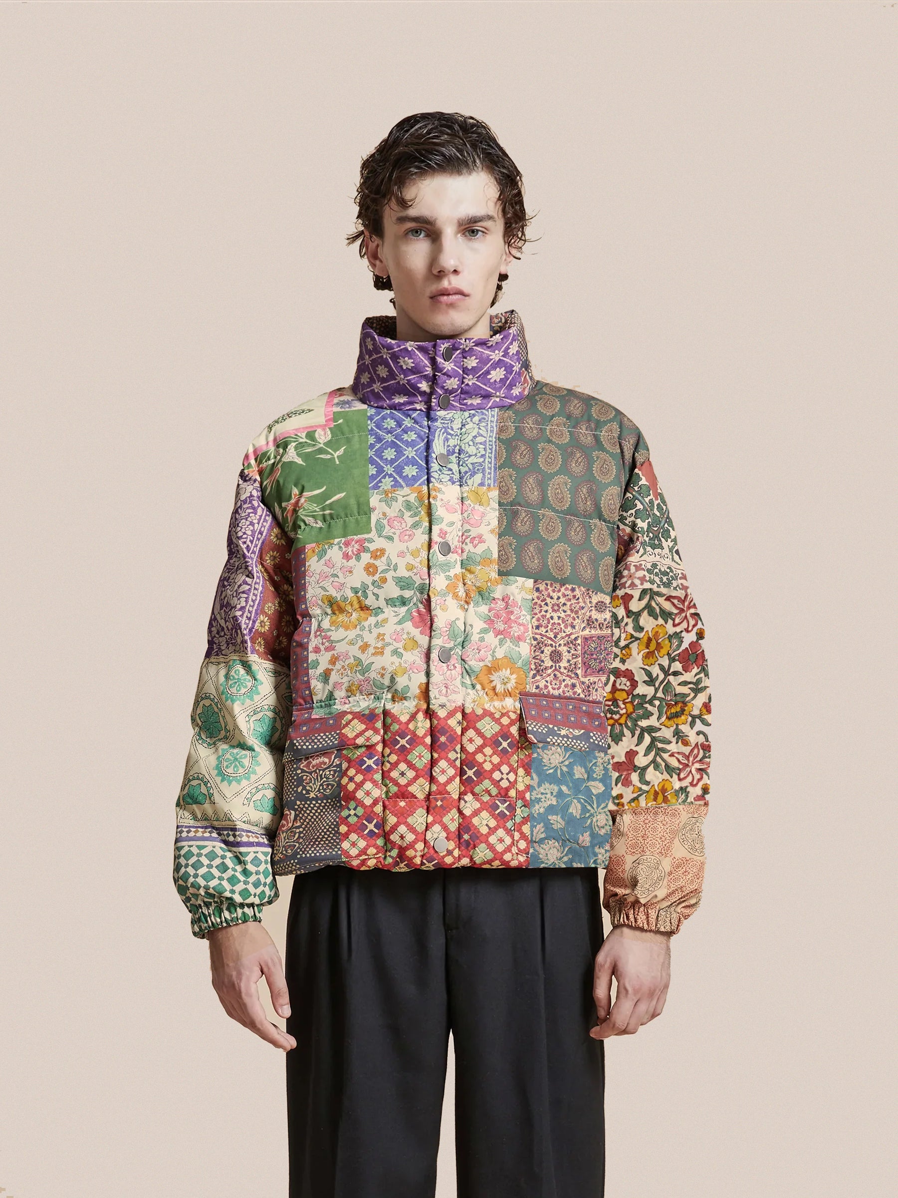 A person wearing a vibrant Gardenia Tapestry Puffer Jacket by Found and black pants stands against a plain background, highlighting the vivid allure of traditional South Asian prints.