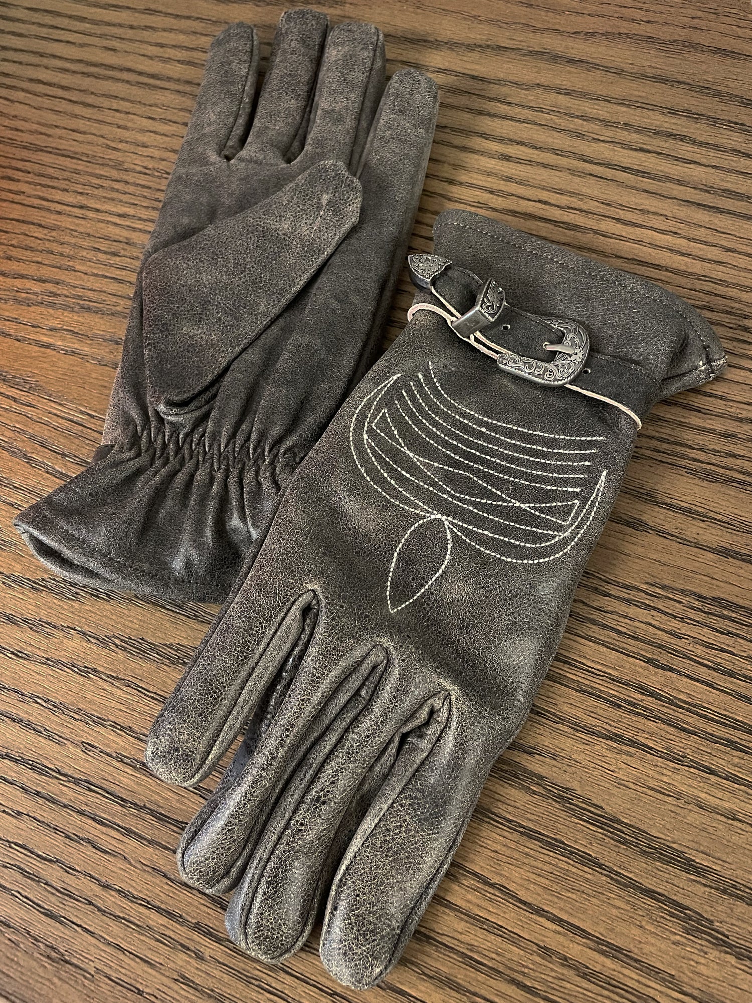 A pair of FOUND's Distressed Leather Western Gloves, featuring stitched detailing and a buckle, placed on a wooden surface.