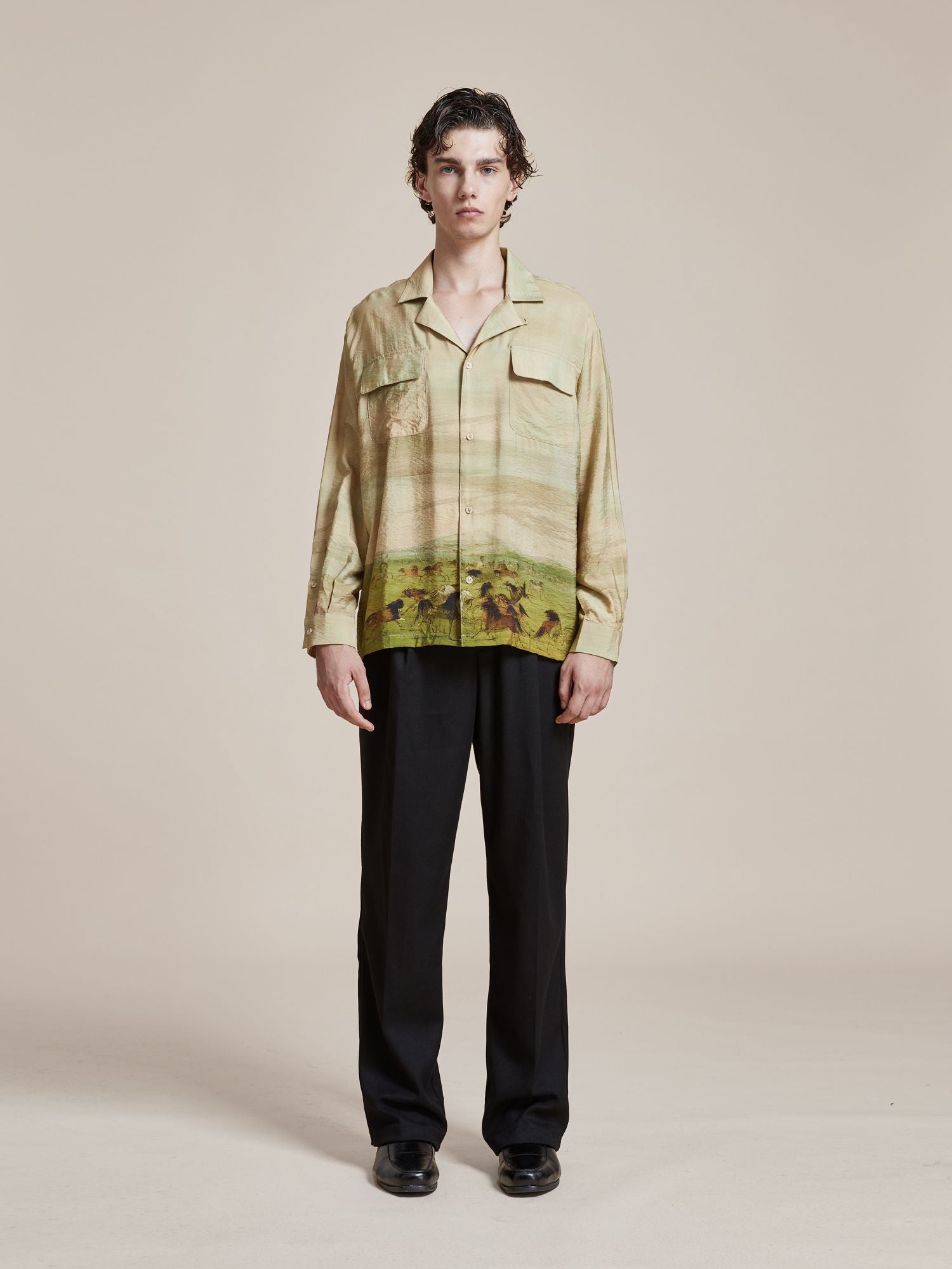 A person with short, curly hair stands facing the camera, wearing a Found Grasslands Long Sleeve Camp Shirt adorned with Phulkari motifs from Punjab Pakistan and black pants against a plain background.