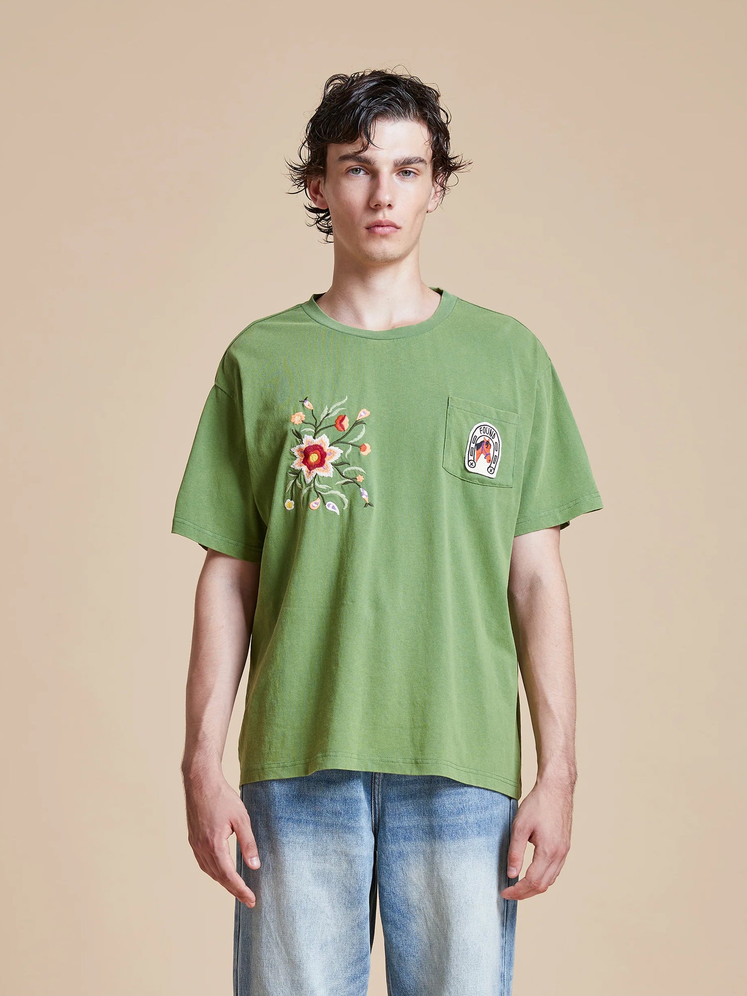 A man wearing a green Found Pine Needle Farm Tee and jeans.