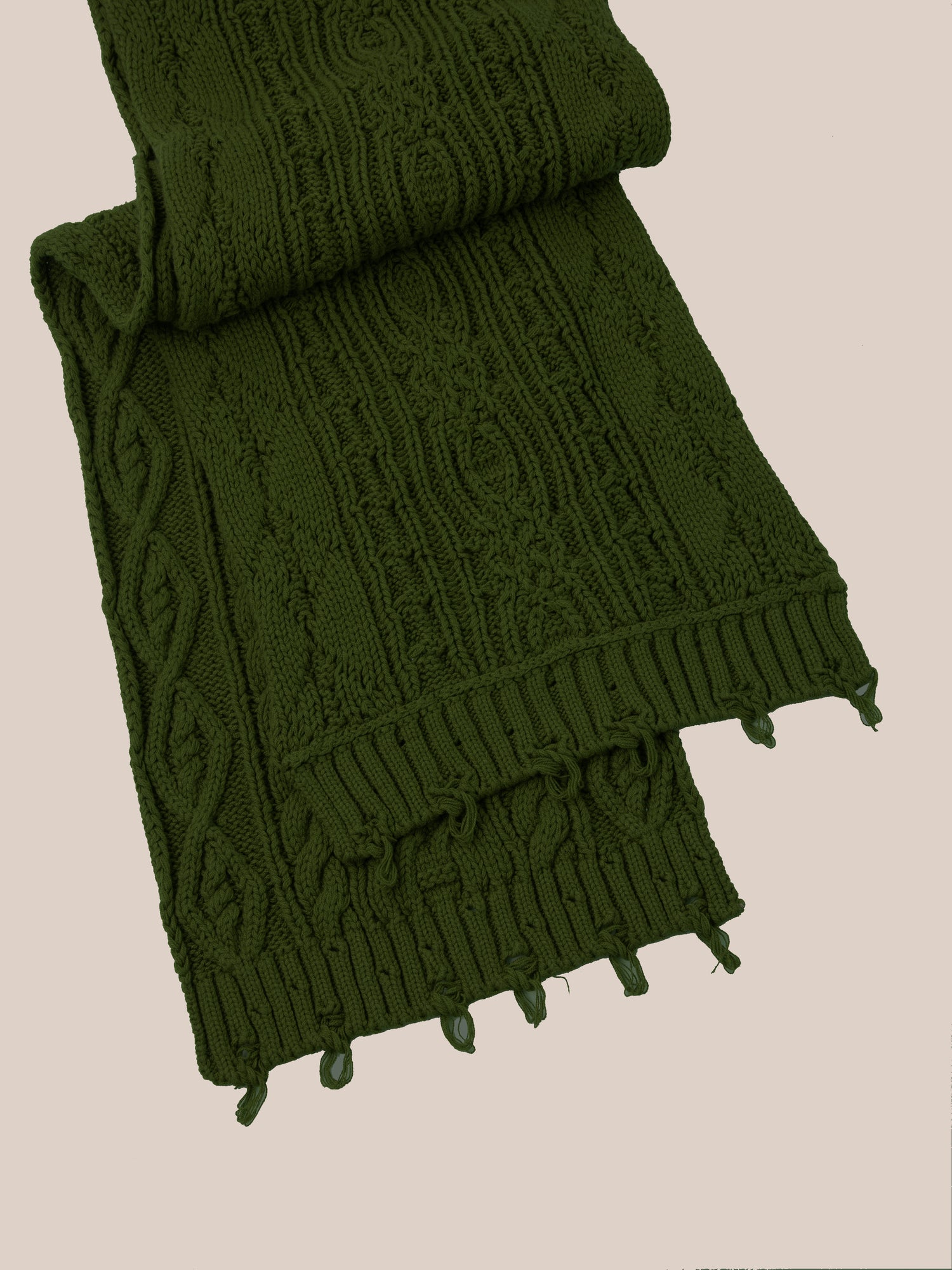 The Forest Distressed Cable Knit Scarf by FOUND, displaying intricate patterns and fringe ends in a folded, dark green knit design, rests against a plain beige background.