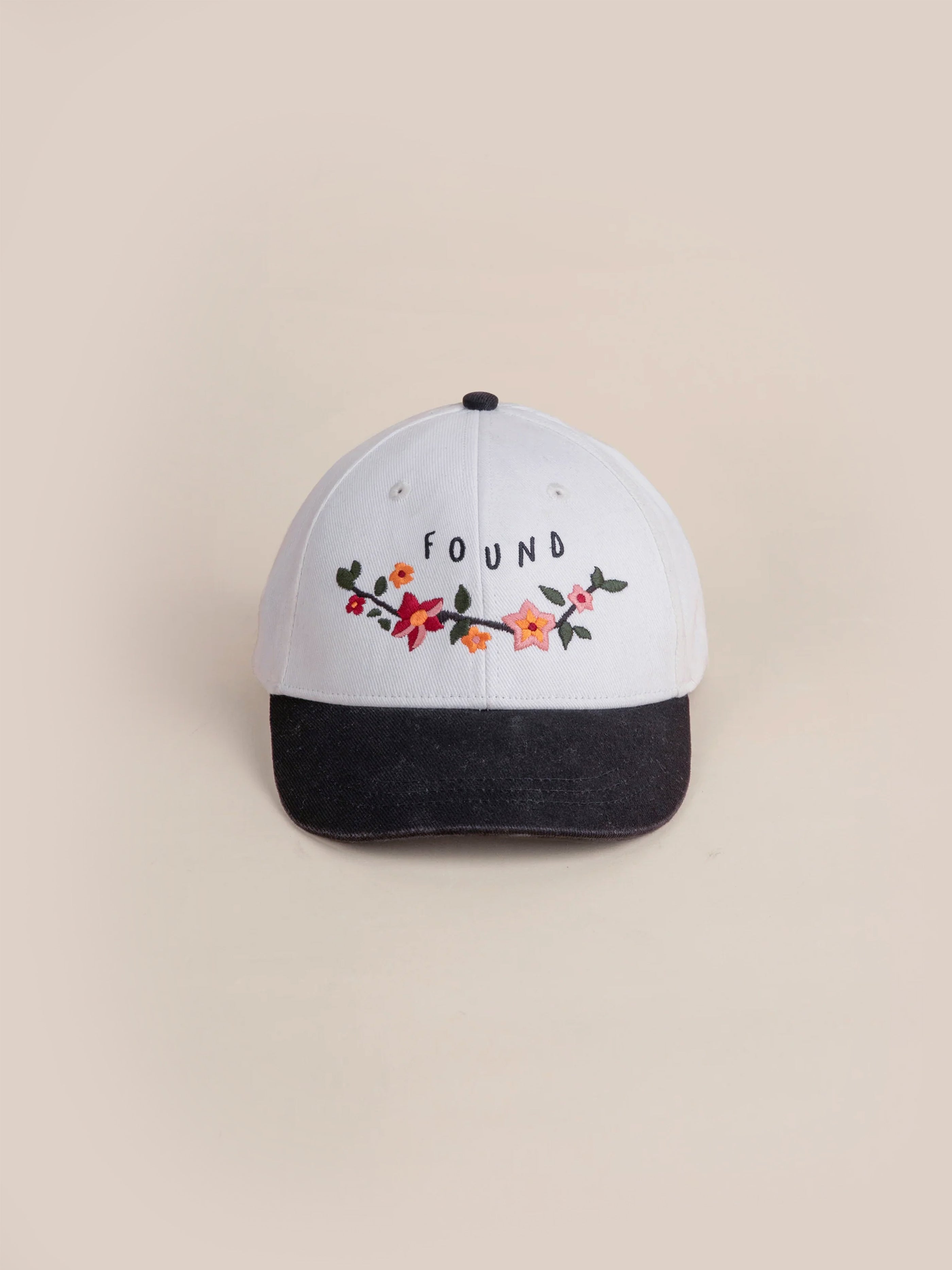 The Greenlands Contrast Cap from FOUND is a white and black cotton cap featuring floral embroidery inspired by Kantha-style artistry, with the brand name prominently displayed on the front.