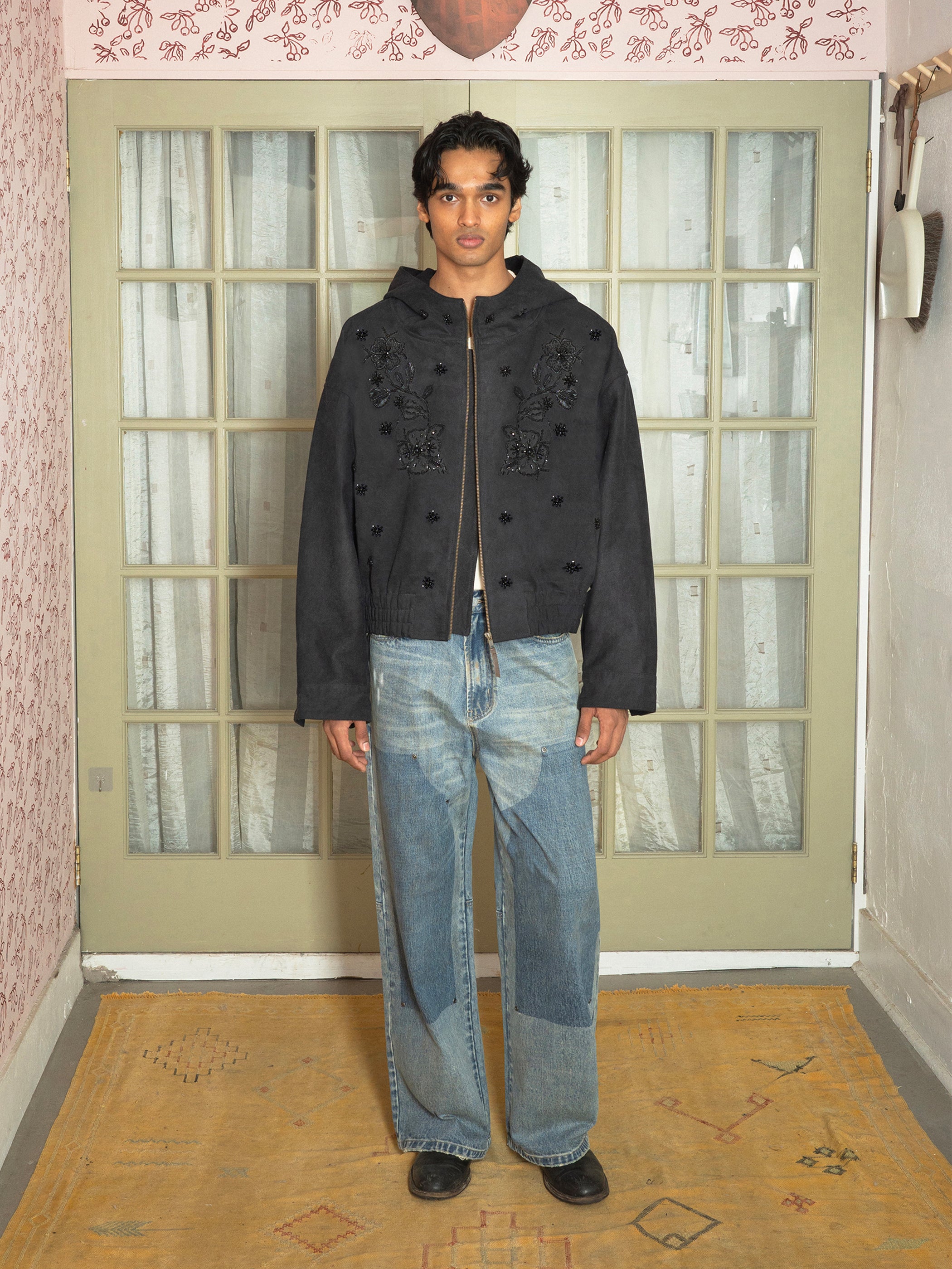 A person stands indoors in the FOUND Hand Embroidered Tonal Anorak Jacket, blue wide-leg jeans, and black shoes, posed in front of glass-panel double doors.
